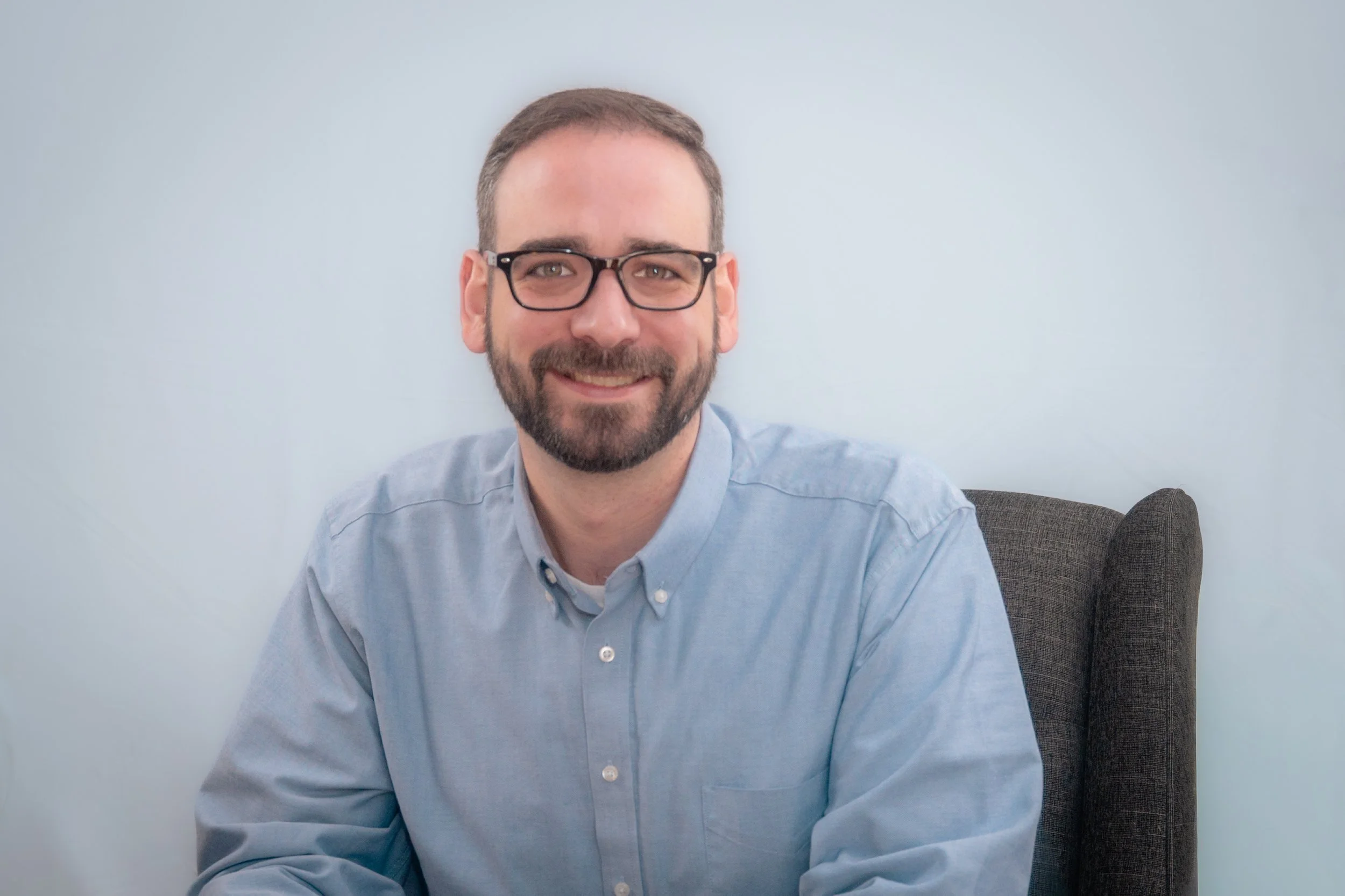 A smiling man with glasses and a beard, sitting on a gray chair against a plain light blue wall, wearing a light blue button-up shirt.