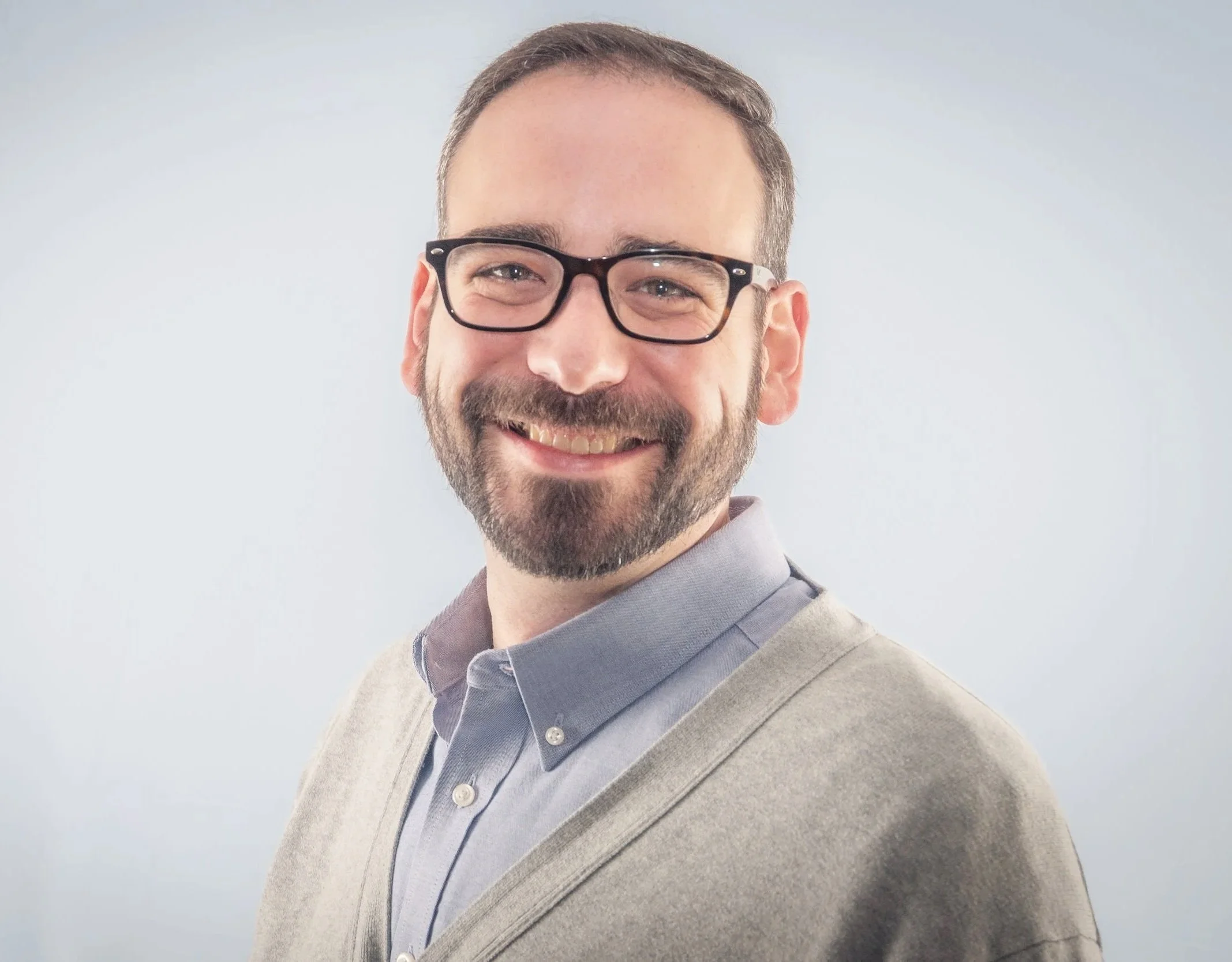 A headshot of a smiling man with glasses, wearing a blue shirt and light gray sweater, against a plain light background.