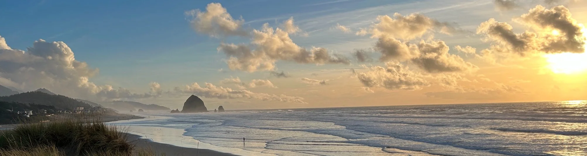 Beach with sand dunes and grasses in the foreground, ocean waves crashing onto the shore, rock formations in the distance, and a setting sun with orange and yellow hues, partly cloudy sky.