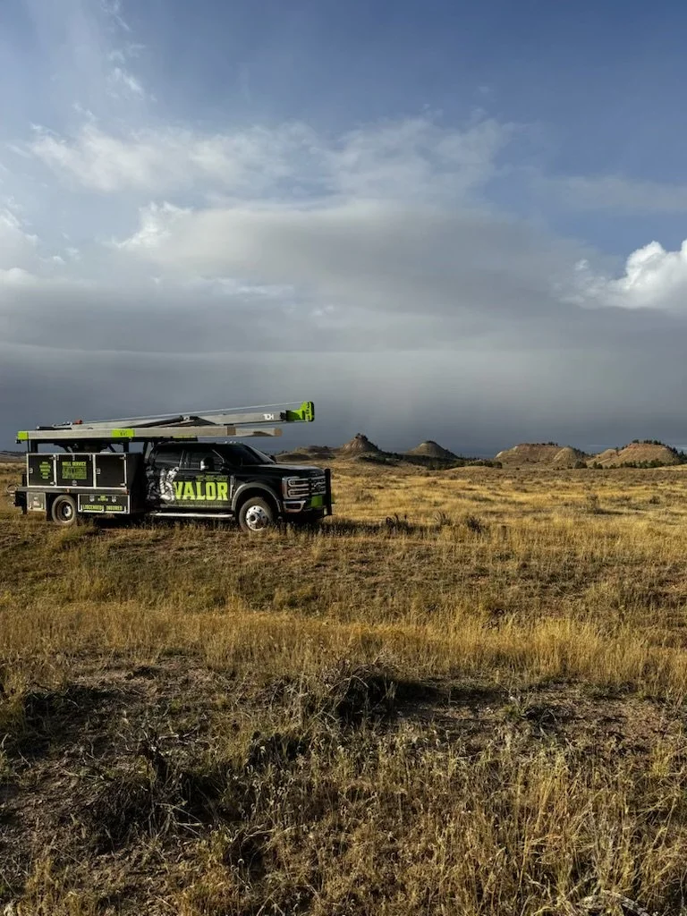A black utility truck with the word 'VALOR' on the side parked in a grassy field under a cloudy sky, with small hills in the background.