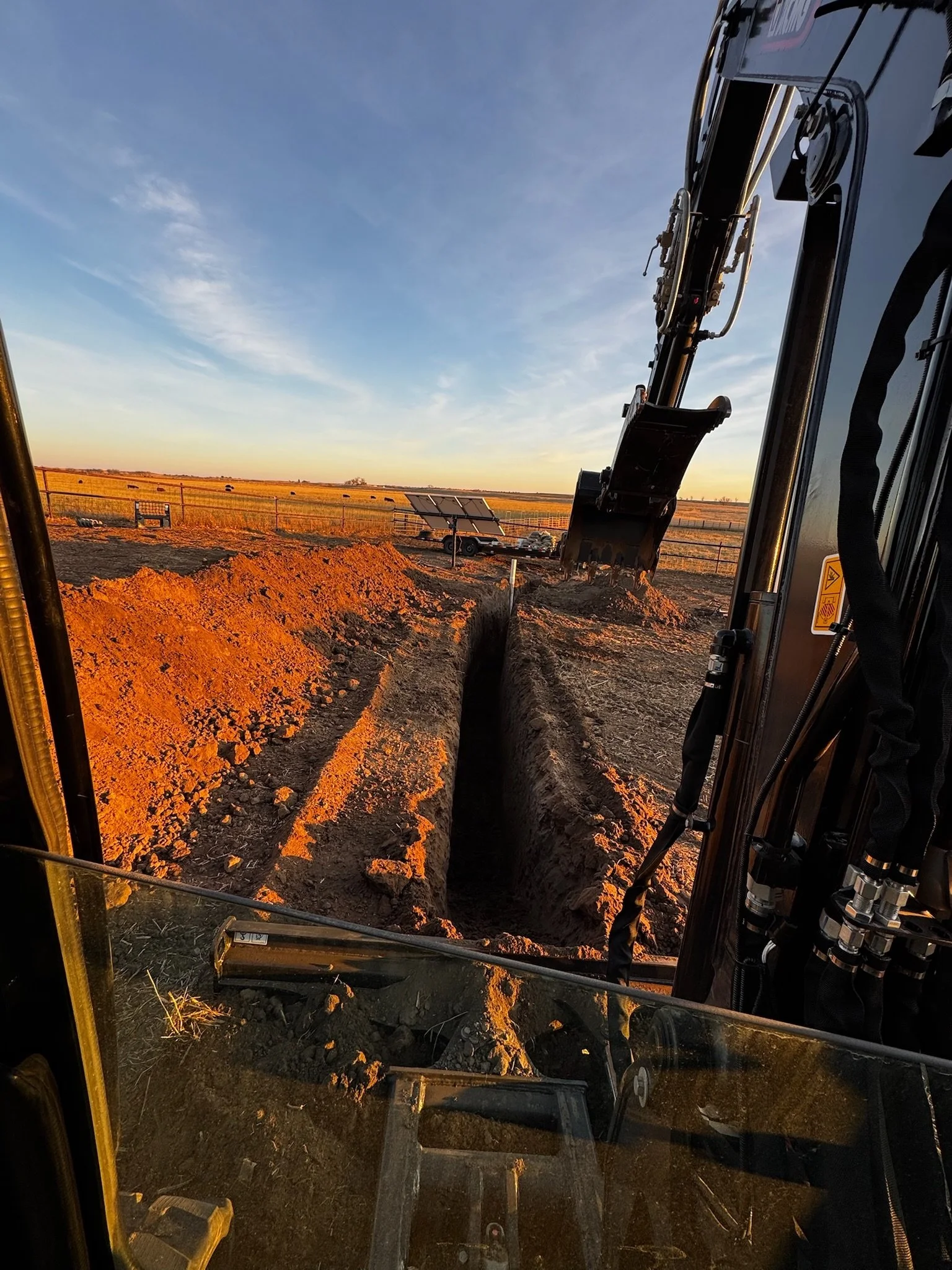 View from the cab of construction equipment looking out at a trench being dug in a dirt field during sunset, with a fence and solar panels in the background.