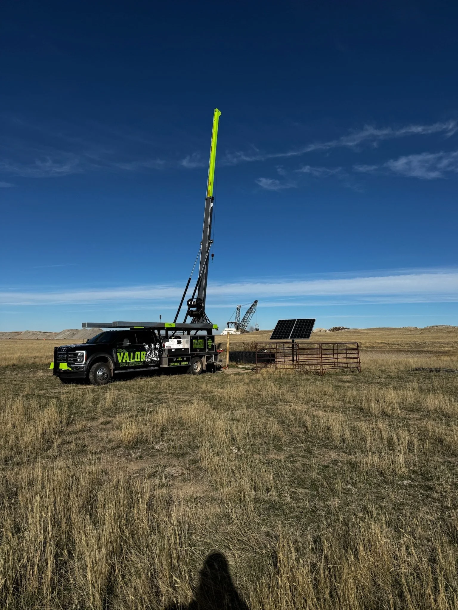 A large black vehicle with green lettering labeled VALOR parked in a field, with a tall surveying instrument extended upward, solar panels and cranes in the background under a blue sky with scattered clouds.