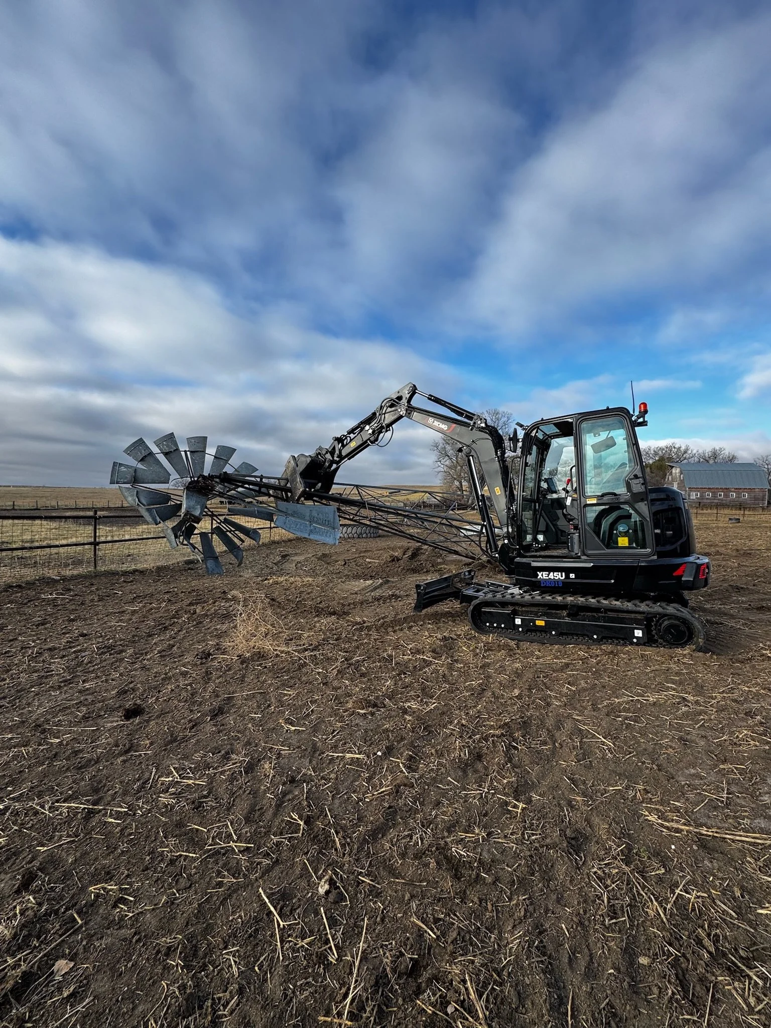 A small black excavator with a rotating arm and a metal attachment working on tilled soil in a rural farm field under a cloudy sky.