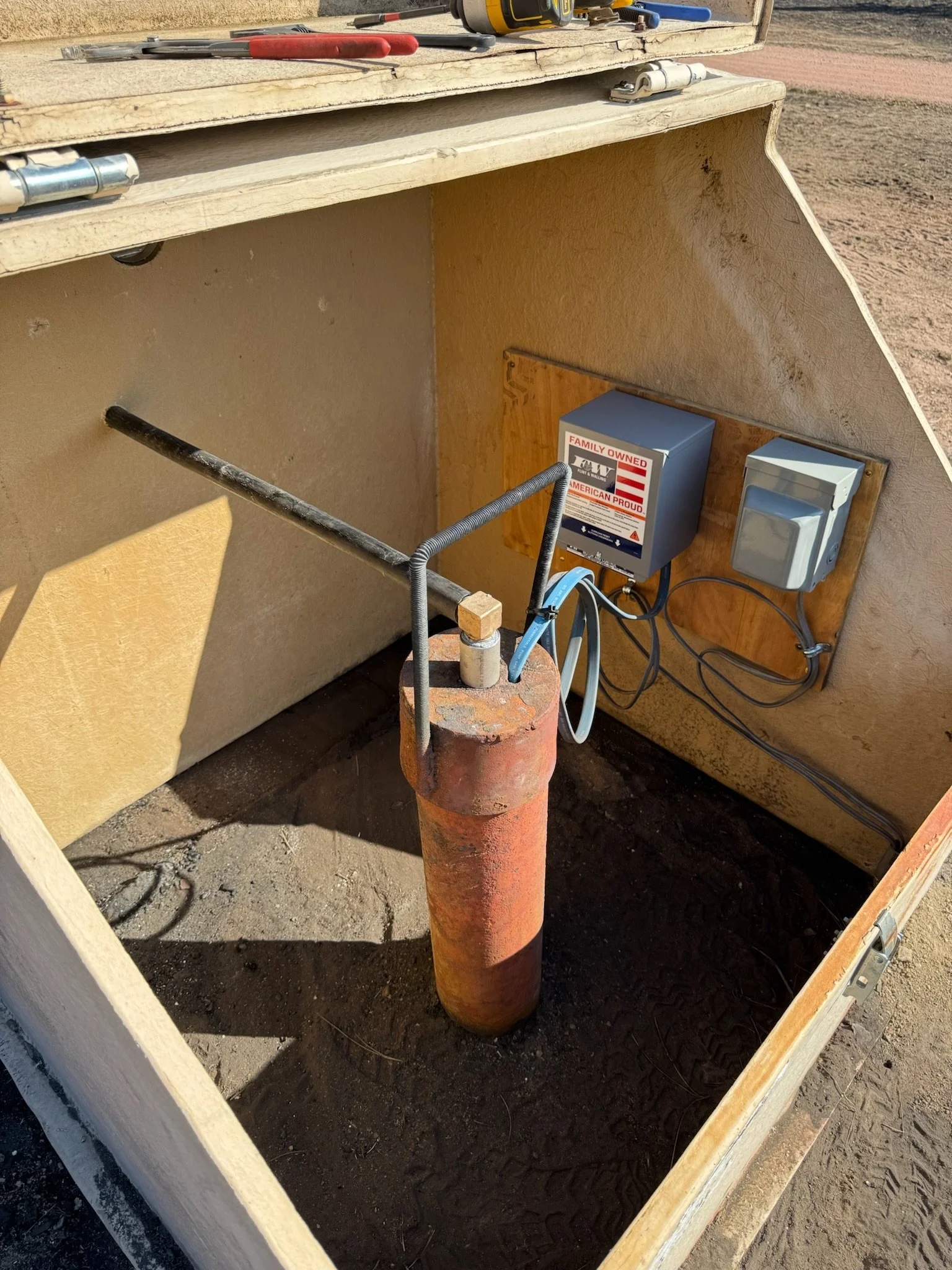 A brick pipe with a metal pipe connection, supporting a metal pipe with wires attached. It is next to a small wooden structure with electrical components, including a utility box with a label that reads "Family Owned, Proud American" and some wiring.