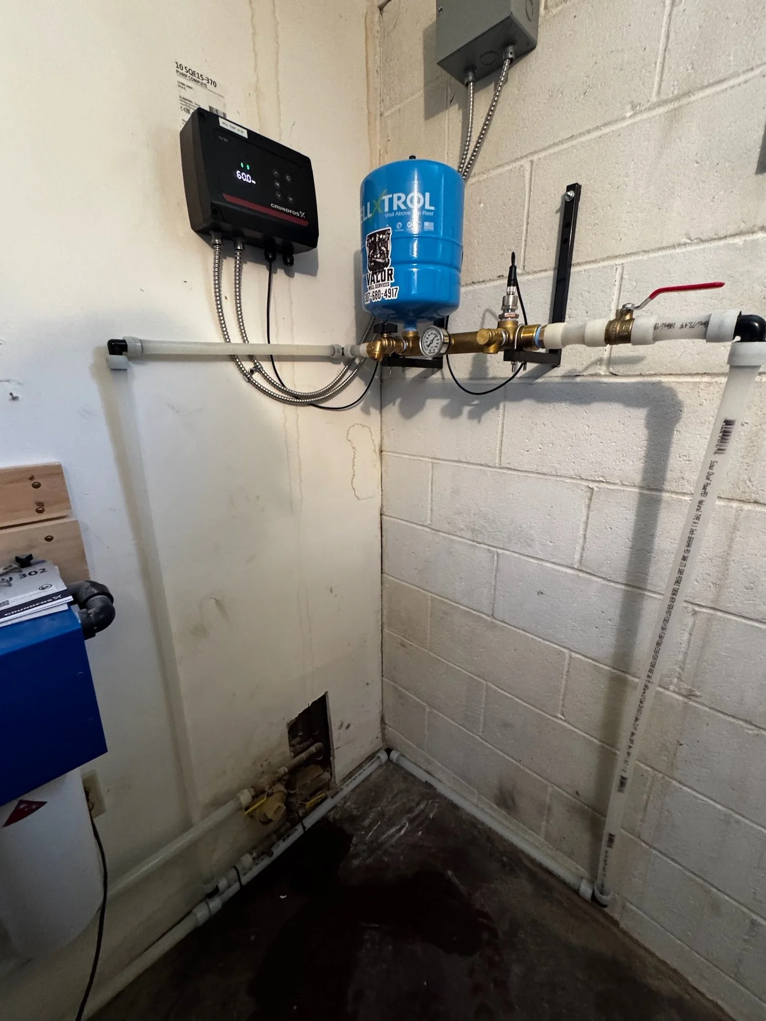 Wall with plumbing and equipment, including a blue pressure tank, digital control panel, gauges, and various pipes in a utility or basement area.