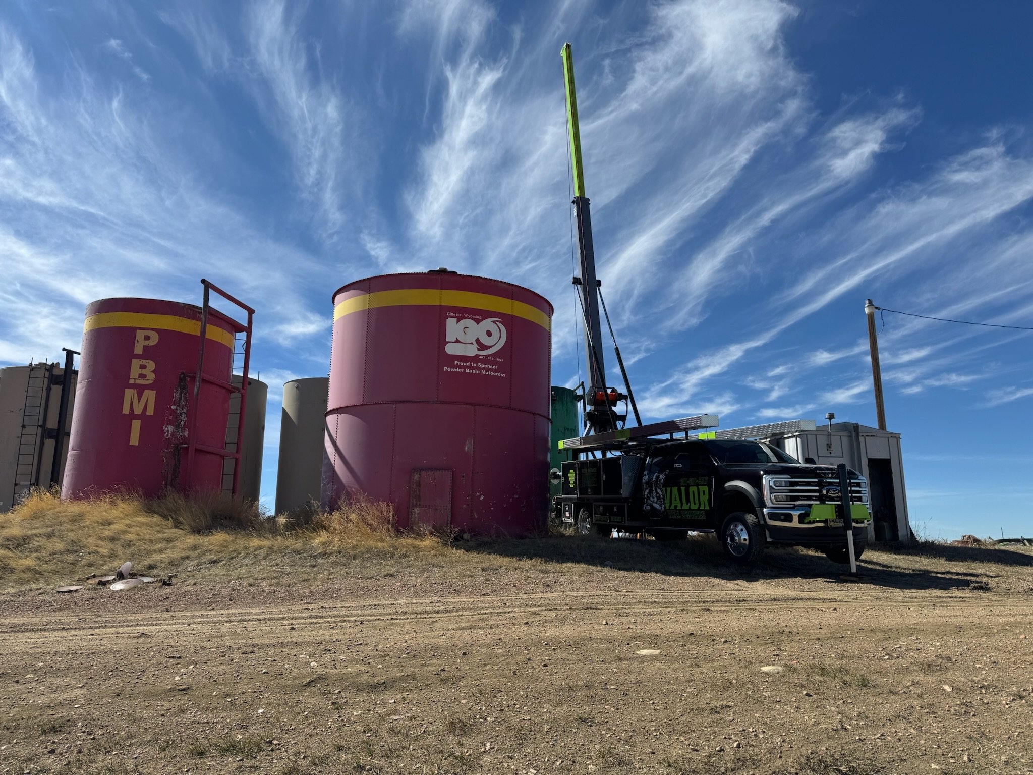 Large pink and yellow tanks labeled 'PBM' and 'KO' standing on a dirt ground under a blue sky with wispy clouds. A black truck with green lettering labeled 'VALOR' is nearby, and a crane extends vertically.