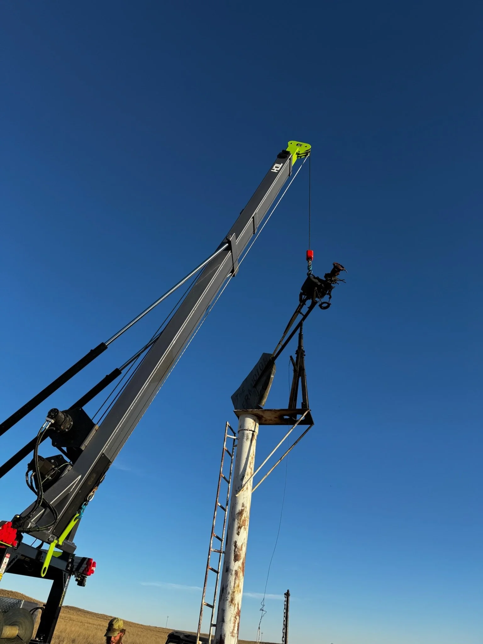 A crane lifting a meteorological instrument, such as a weather monitoring device, against a clear blue sky, with a field in the background.