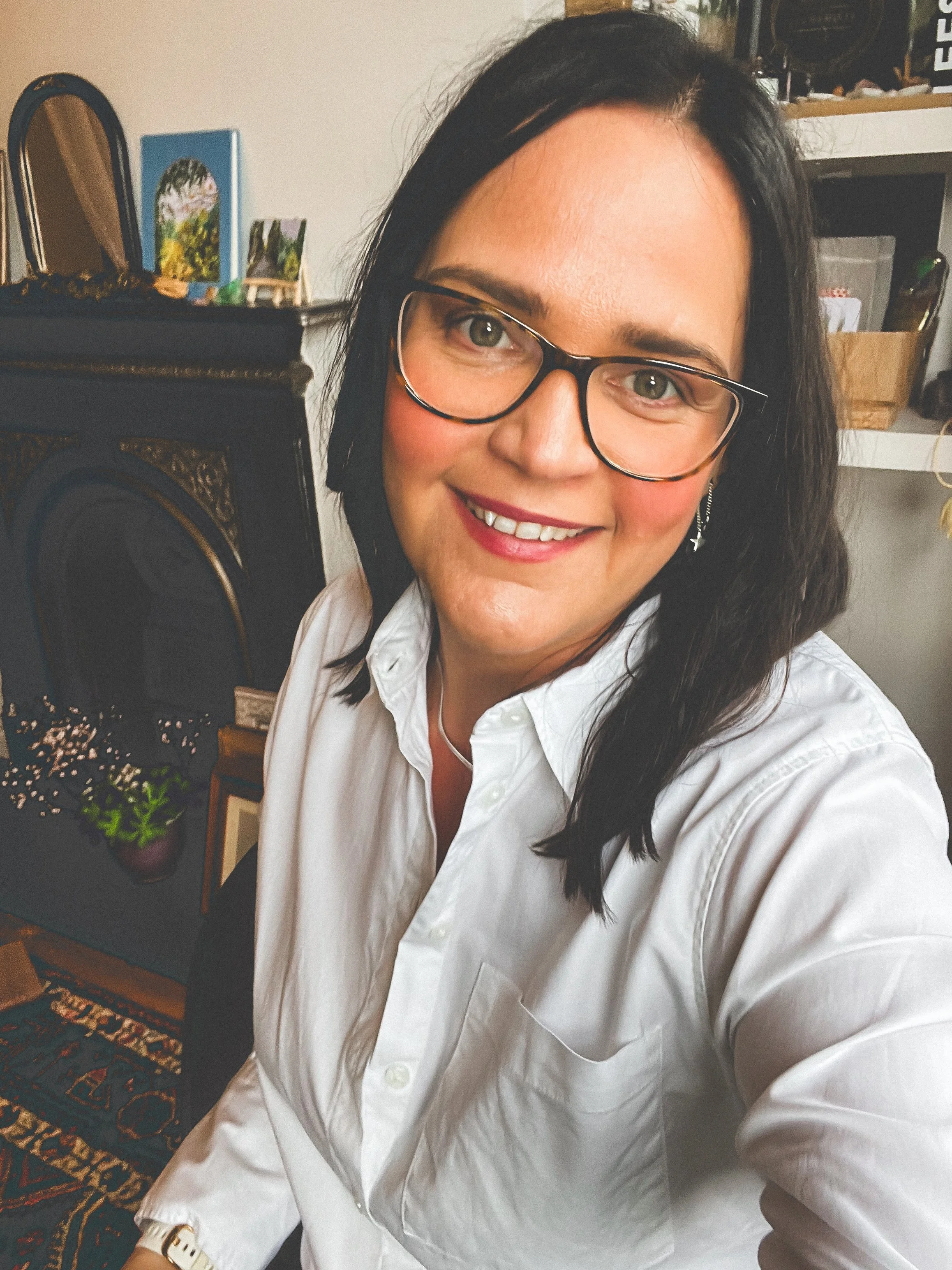 A smiling woman with glasses and dark hair taking a selfie indoors, wearing a white button-up shirt.