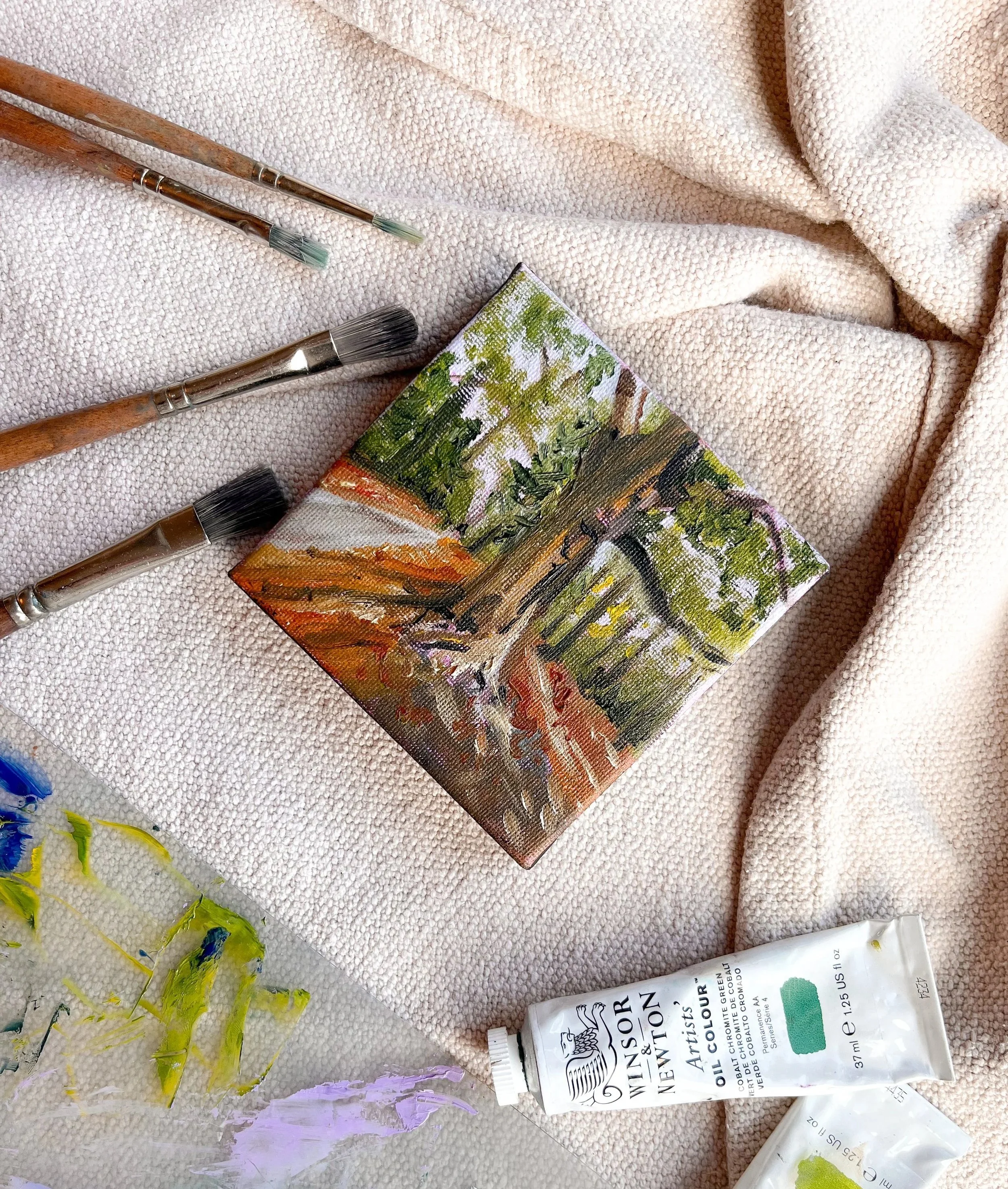Paintbrushes, an empty painted canvas with trees, and paint tubes on a textured fabric surface, indicating an art or painting scene.