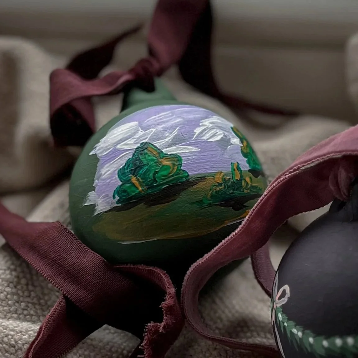 Bespoke hand-painted Christmas bauble featuring a miniature green landscape painting, finished with a burgundy ribbon.