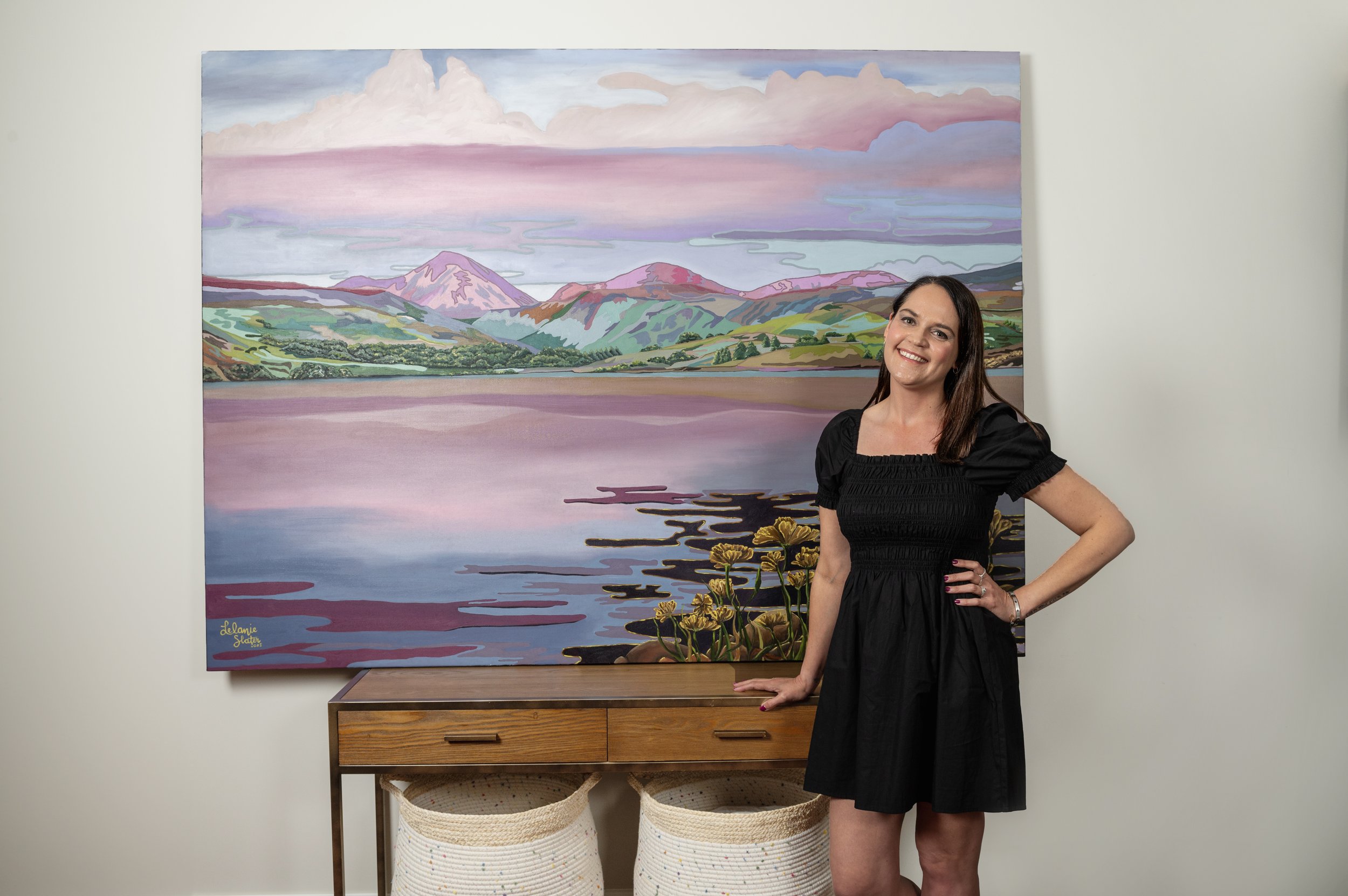 A woman with long dark hair in a black dress smiling and standing next to a landscape painting of mountains, water, and sky in pastel colors, in an art gallery or home setting.