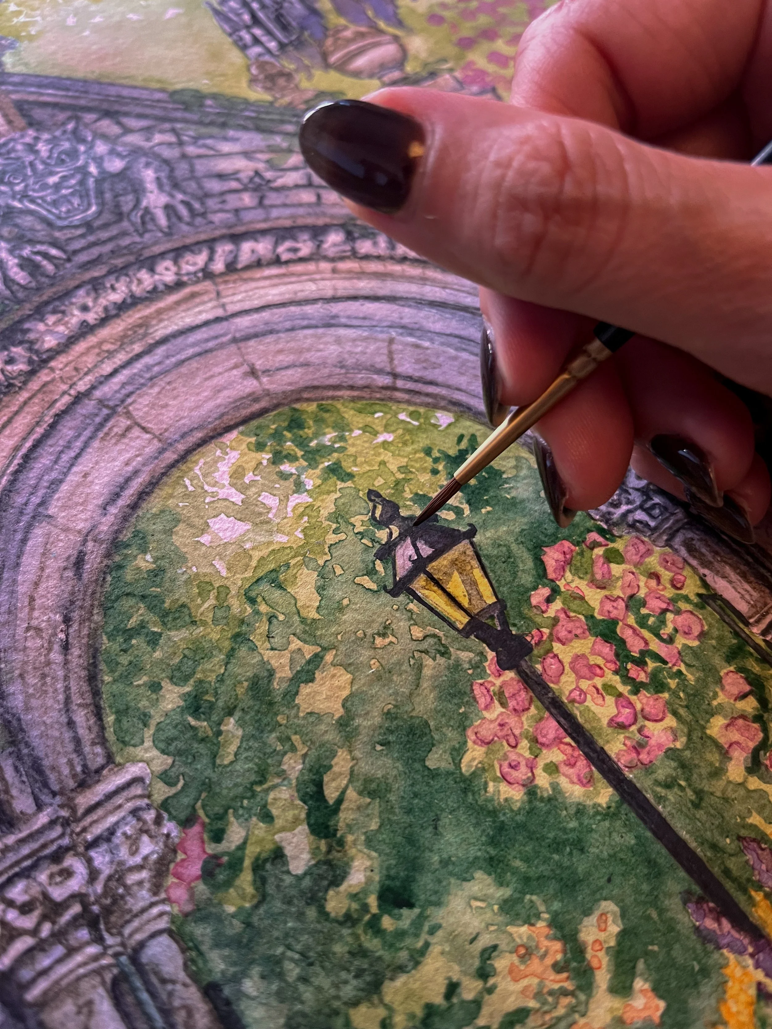 Close-up of a hand with dark nail polish holding a fine-tipped paintbrush, painting a detailed scene of an archway, a lantern, and lush greenery with pink flowers in a watercolor style.