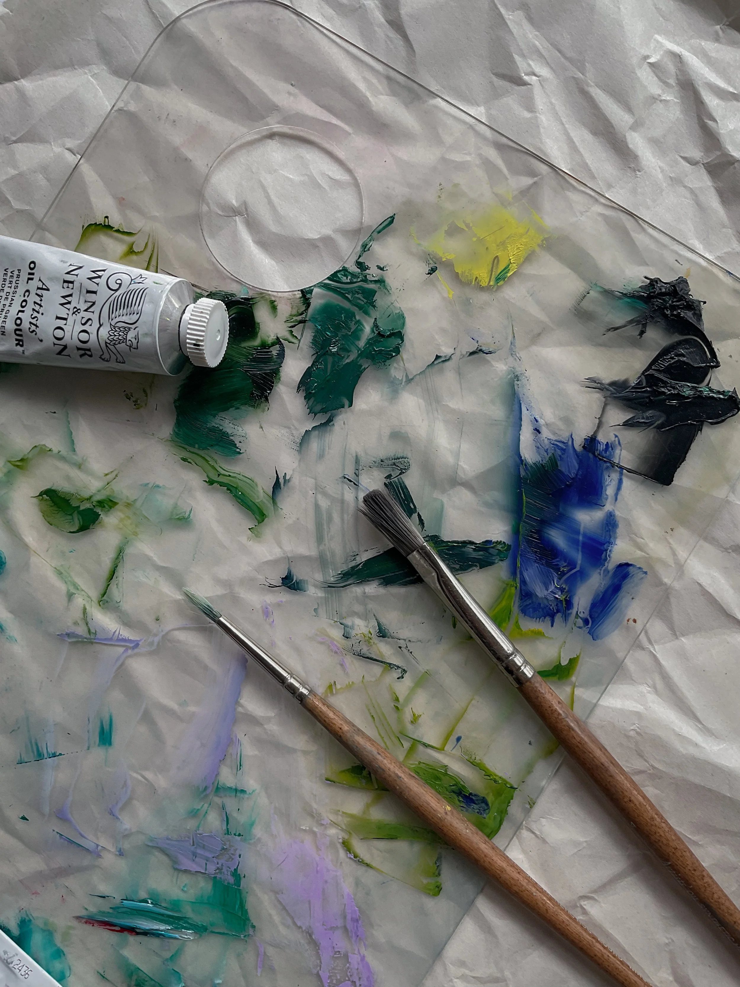 A crumpled sheet of white paper covered with multicolored paint smudges, a tube of Winsor & Newton artist's oil color, and two paintbrushes with wooden handles.