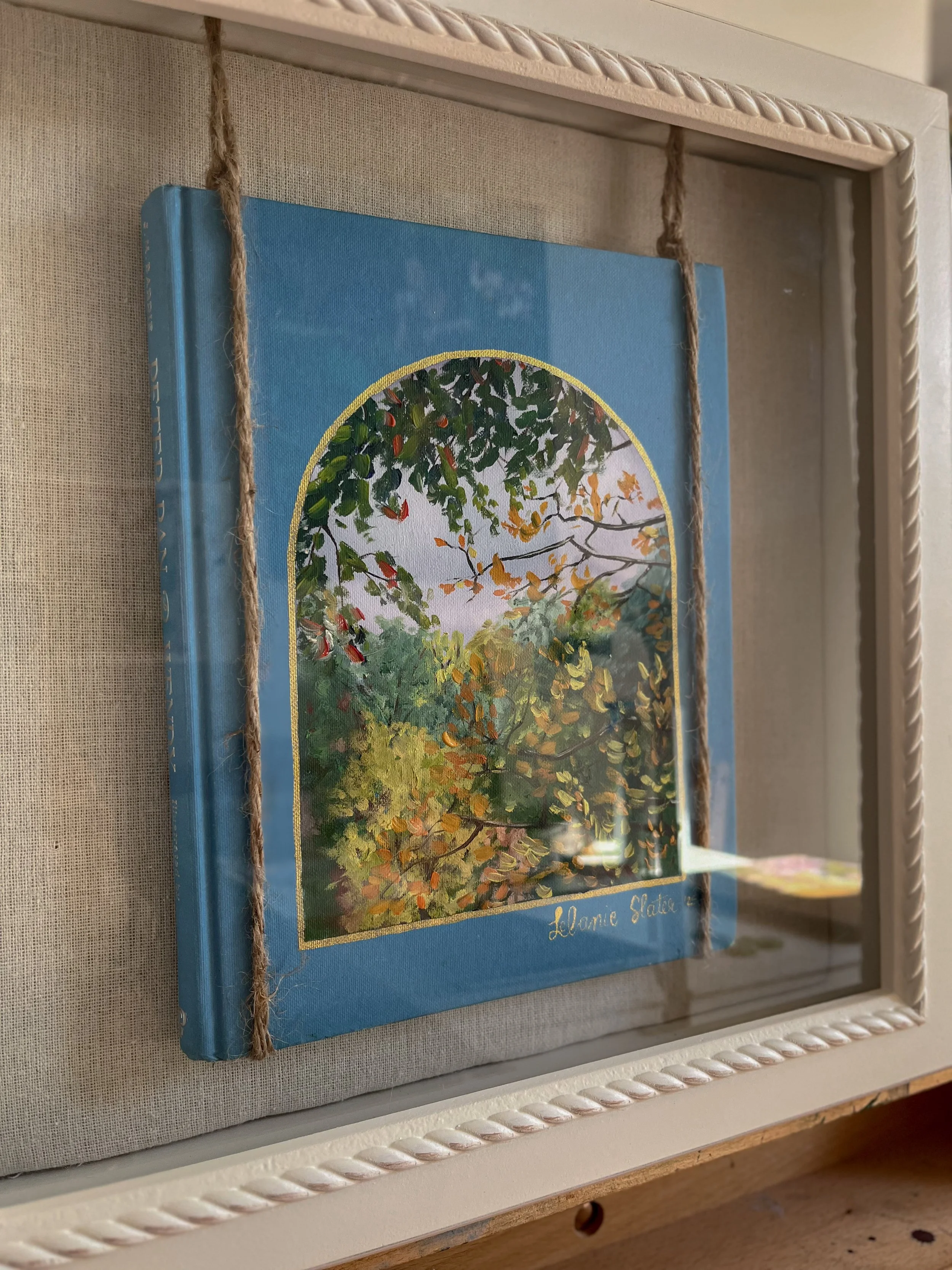A framed picture of a colorful outdoor scene with trees and leaves, with a blue border, inside a white frame with a twisted rope design, on a beige fabric background.