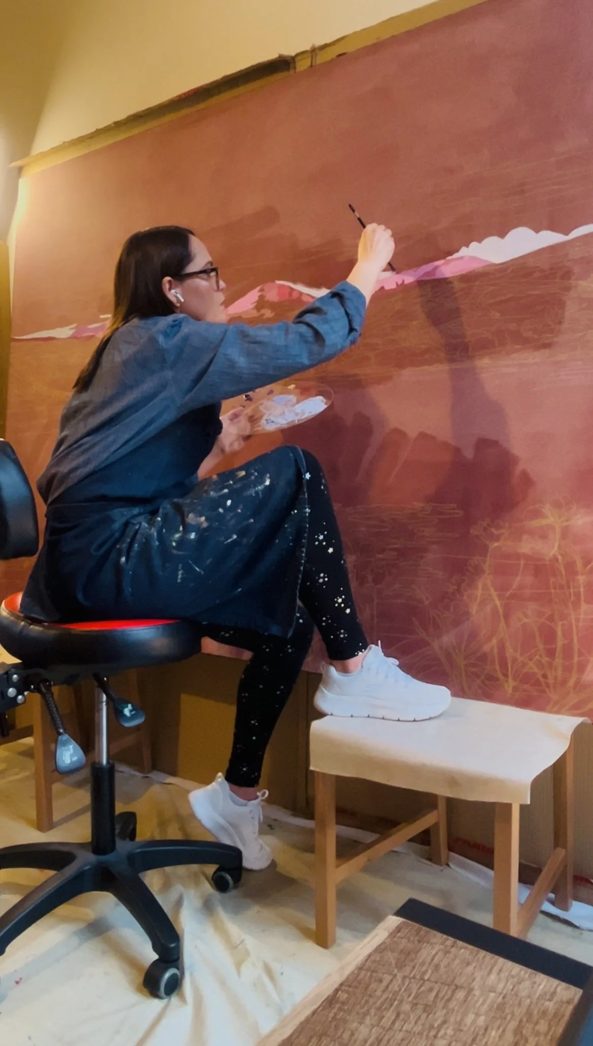 Artist painting a landscape on a wall mural, sitting on a chair and holding a palette and brush, with furniture and a painted background.
