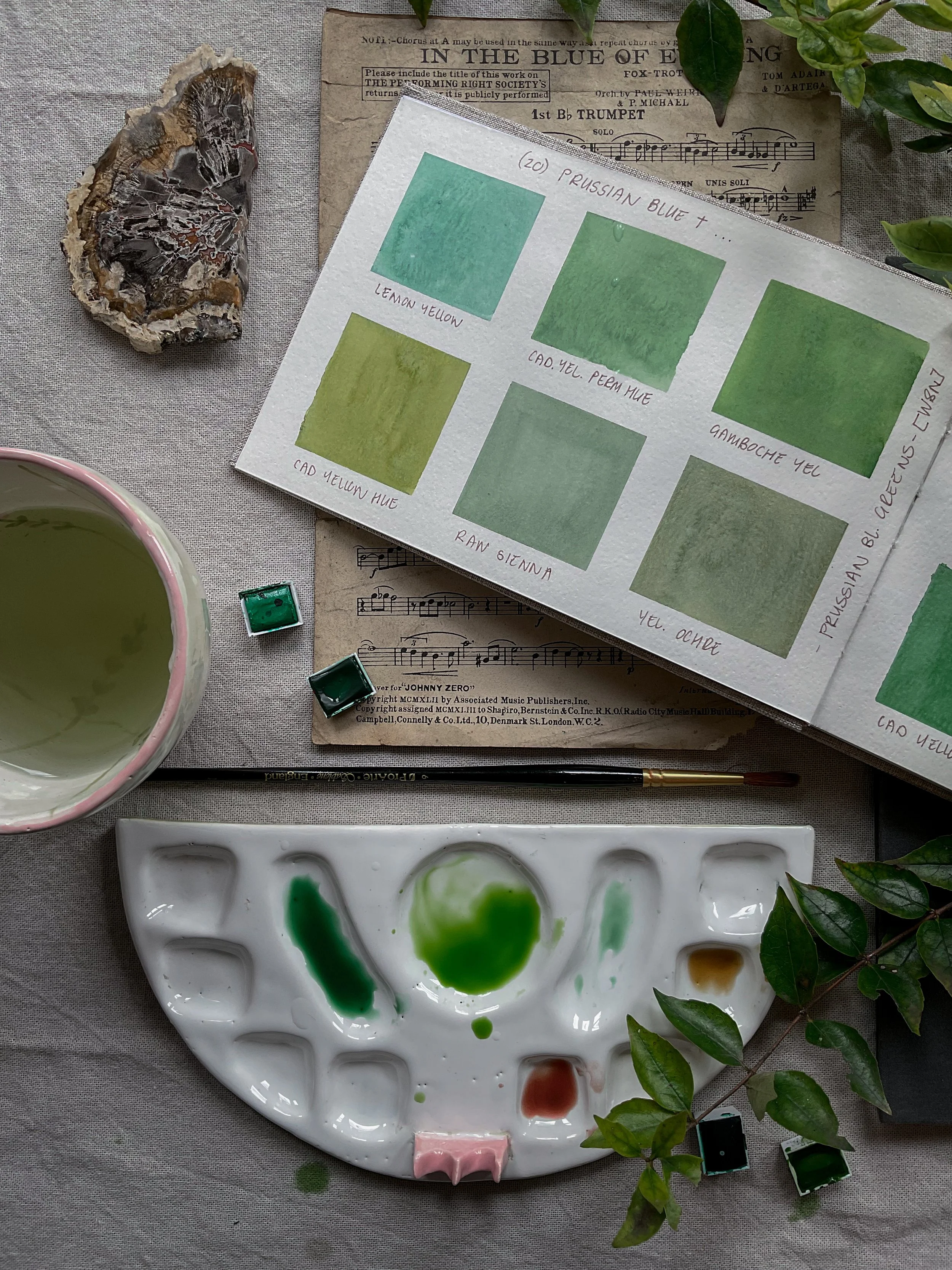 A watercolor paint palette with green shades, a pink and green leaf, a ceramic cup with watercolor paint, a sheet of paper with watercolor swatches, a rock specimen, and musical sheets on a fabric surface.