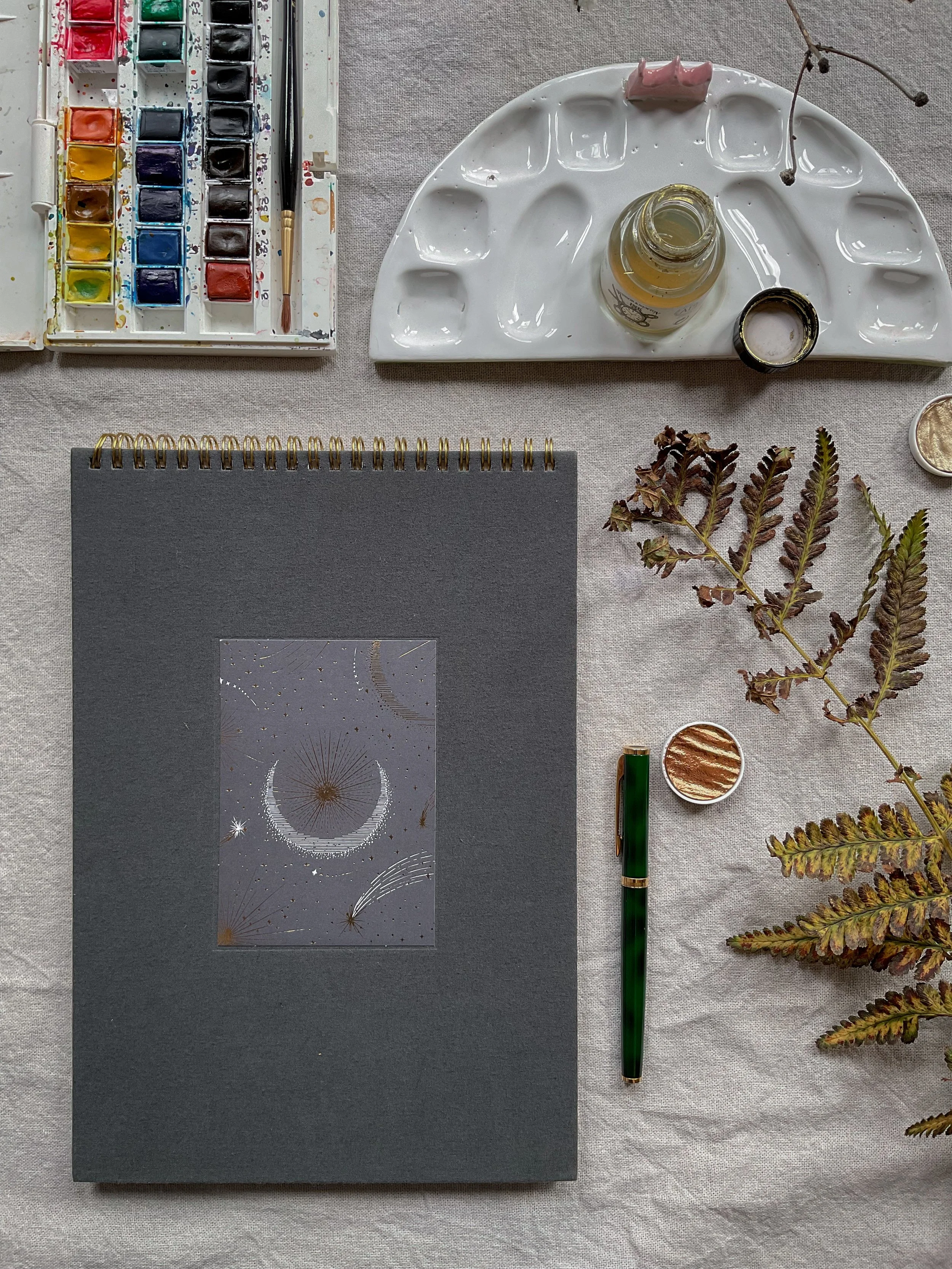 A watercolor paint set, a paintbrush, a bottle of ink, a small container of white paint, a closed notebook or journal, and some dried fern leaves on a light-colored fabric surface.