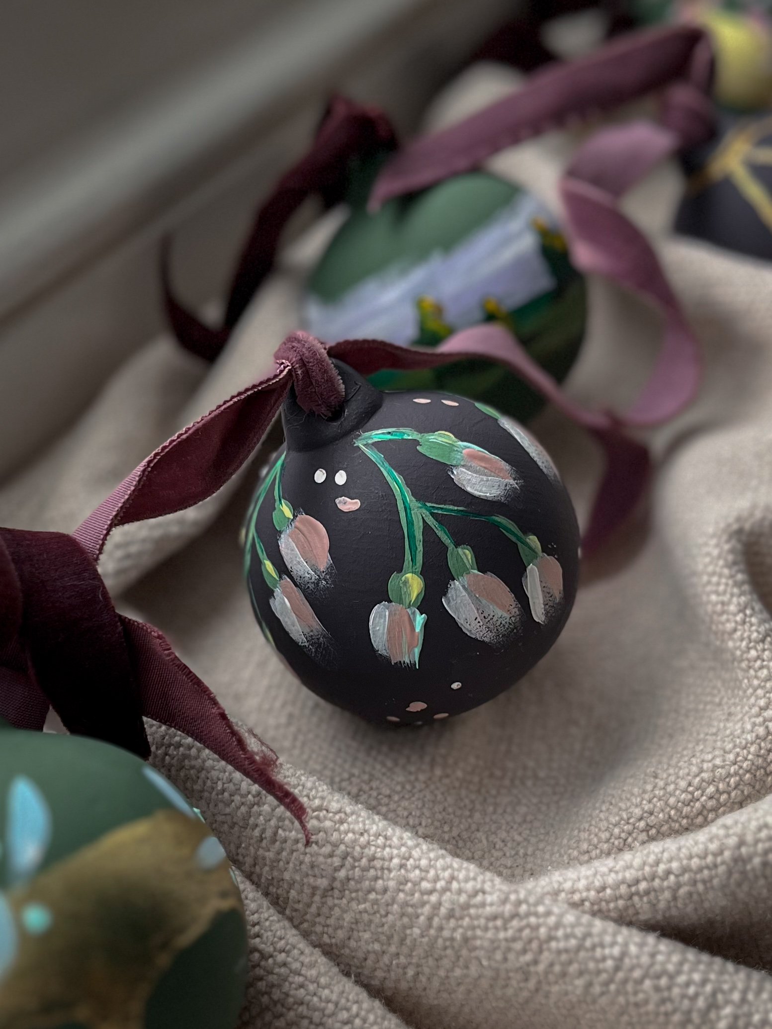 Bespoke hand-painted ceramic Christmas bauble featuring delicate pink, green and white botanical designs on a dark background, finished with a burgundy ribbon.