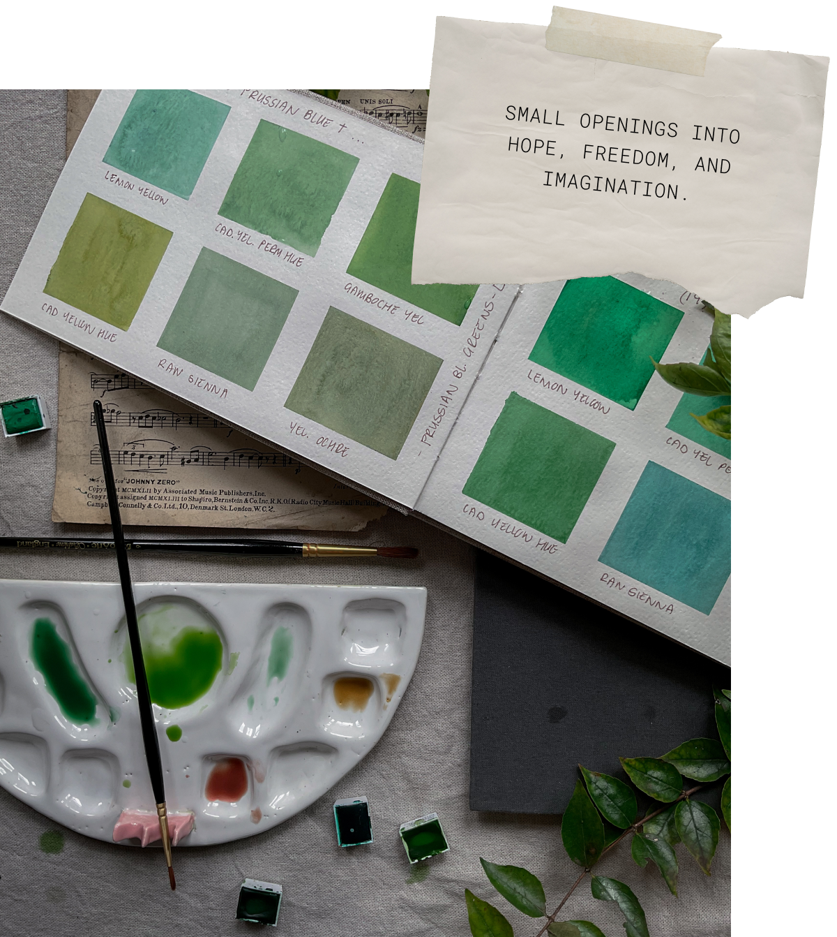 A watercolor palette with green, yellow, and brown paints, paintbrushes, and a sheet of music in the background. An open book displays various shades of green and blue watercolor samples, labeled with names such as 'Lemon Yellow,' 'Cad Yellow Hue,' and 'Raw Sienna.' A torn piece of paper with a motivational quote, 'Small openings into hope, freedom, and imagination,' is taped to the right side of the book. Green leaves are placed nearby as decoration.