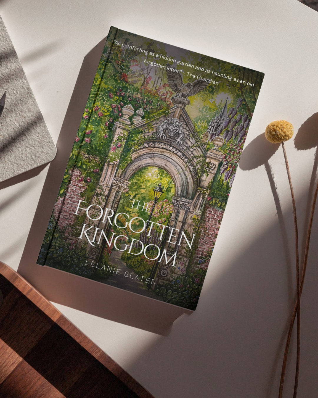 A book titled 'The Forgotten Kingdom' by Lelanie Slater lying on a white surface with a dried flower and a paper with textured edges nearby.
