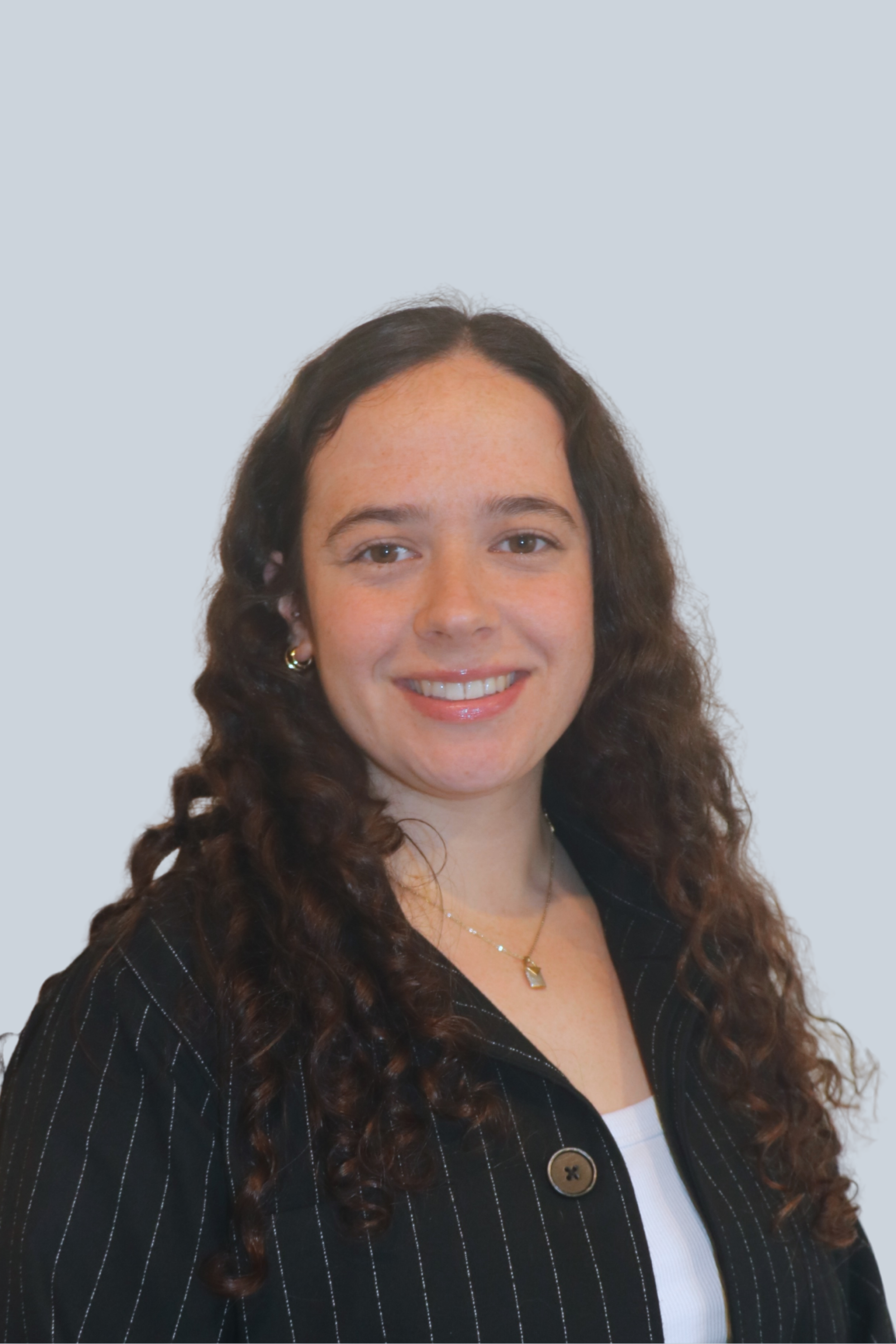 A woman with long curly dark hair, wearing a black pinstripe blazer and a white top, smiling at the camera. She has small earrings and a delicate necklace, standing against a light blue background.