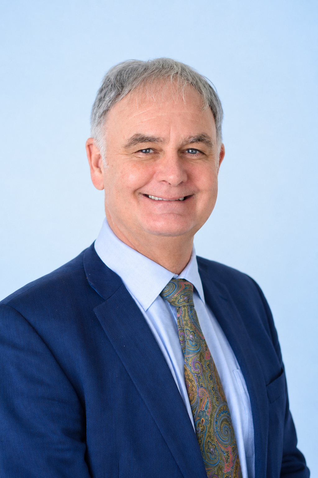 Headshot of a middle-aged man with gray hair, wearing a dark blue suit, light blue shirt, and a colorful paisley tie, smiling against a light blue background.