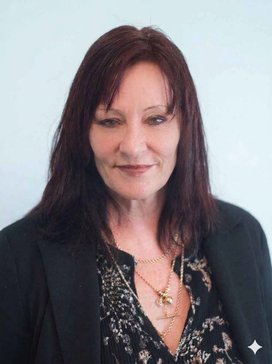 Portrait of a woman with shoulder-length dark red hair, wearing a black blazer over a patterned blouse, and layered gold necklaces, standing against a plain light background.