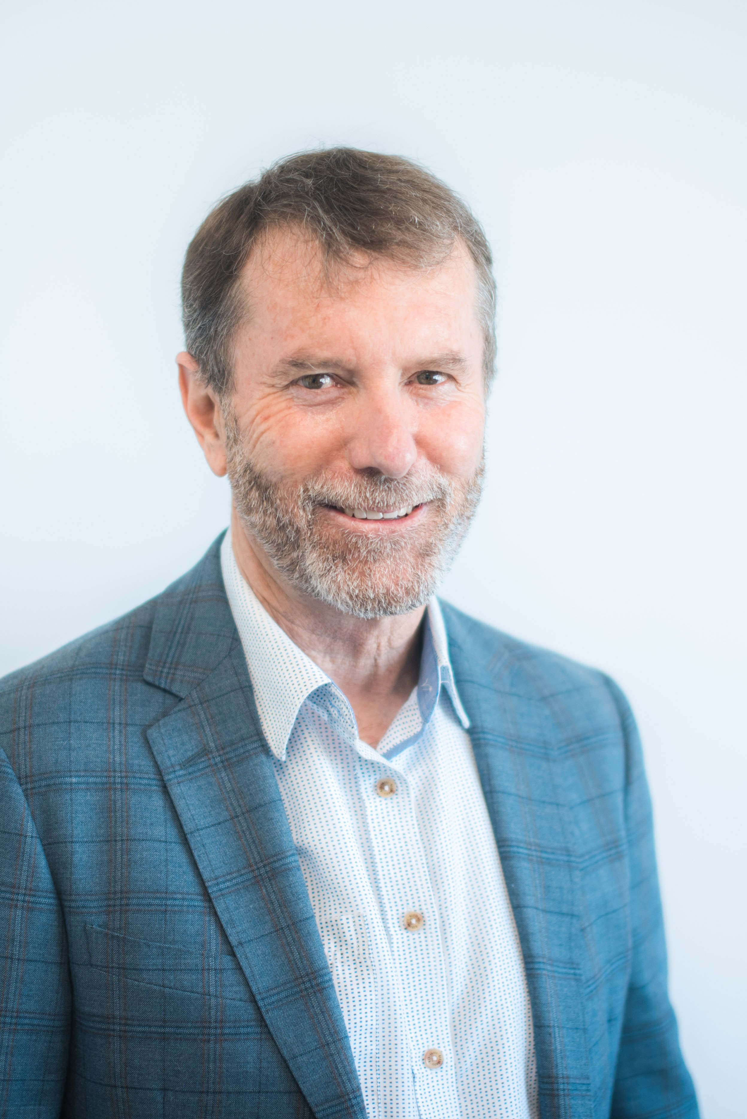 A middle-aged man with gray hair and a beard, wearing a blue checked blazer and a white shirt, smiling against a light blue background.