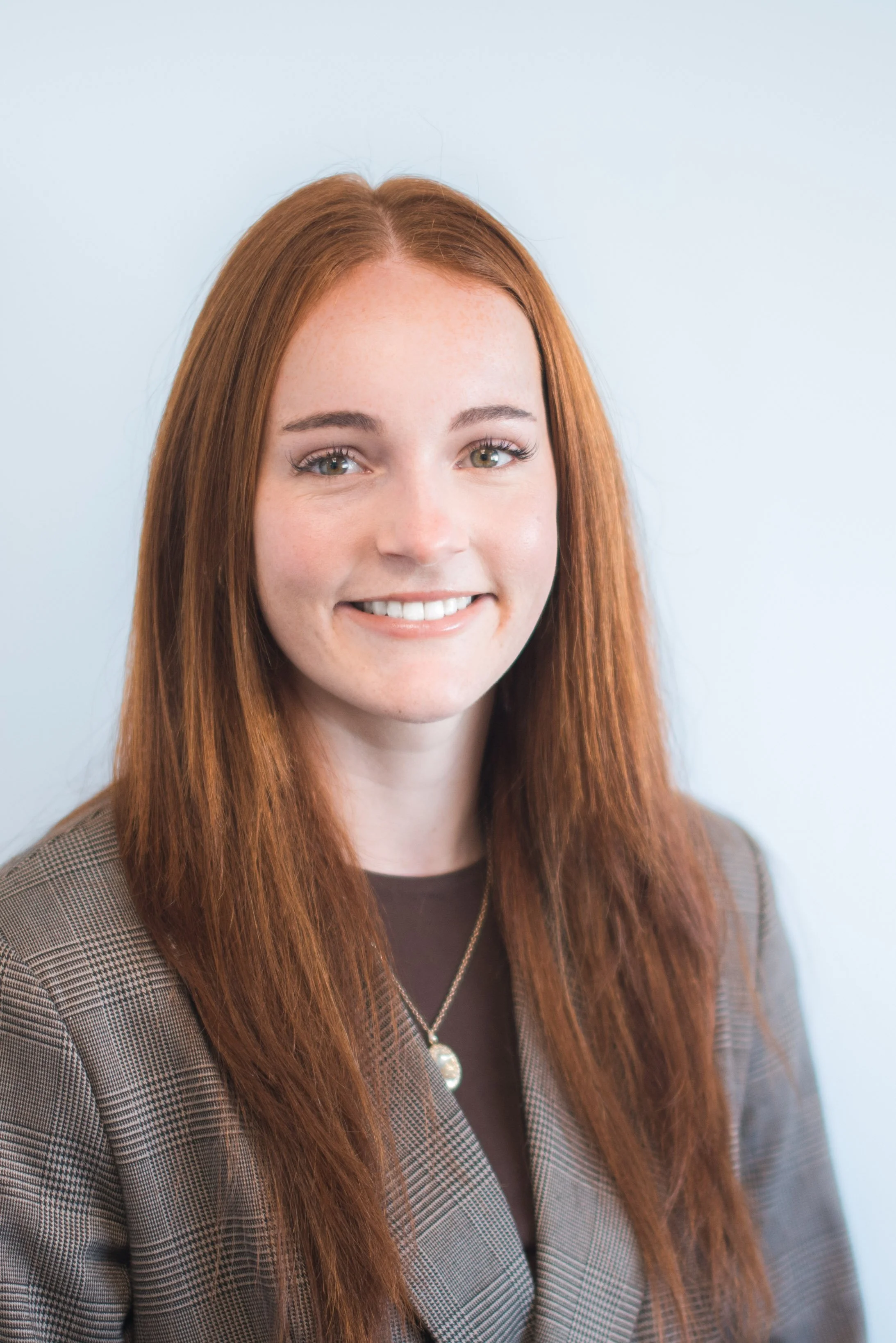 A woman with long red hair and hazel eyes, smiling, wearing a plaid blazer over a dark top, accessorized with a necklace with a pearl pendant, standing against a plain light blue background.