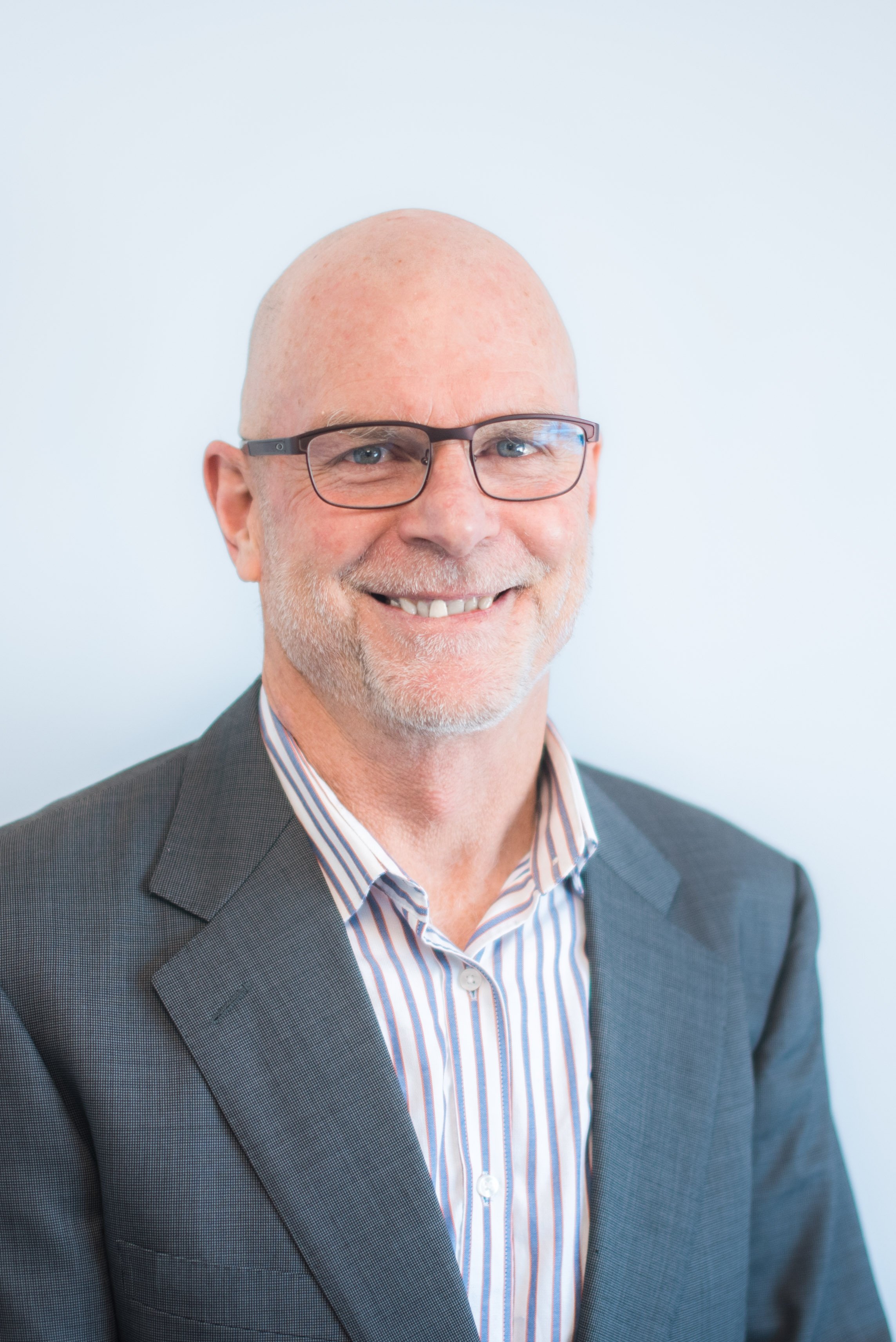 A middle-aged man with a bald head, gray beard, wearing glasses, a dark gray suit, and a striped dress shirt, smiling in front of a plain white background.
