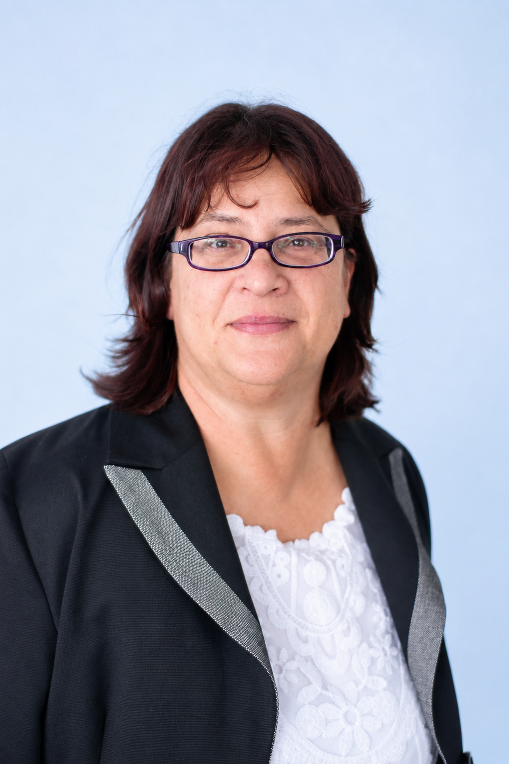 A woman with shoulder-length dark brown hair, wearing eyeglasses, a black blazer with silver trim, and a white embroidered blouse, standing against a light blue background.