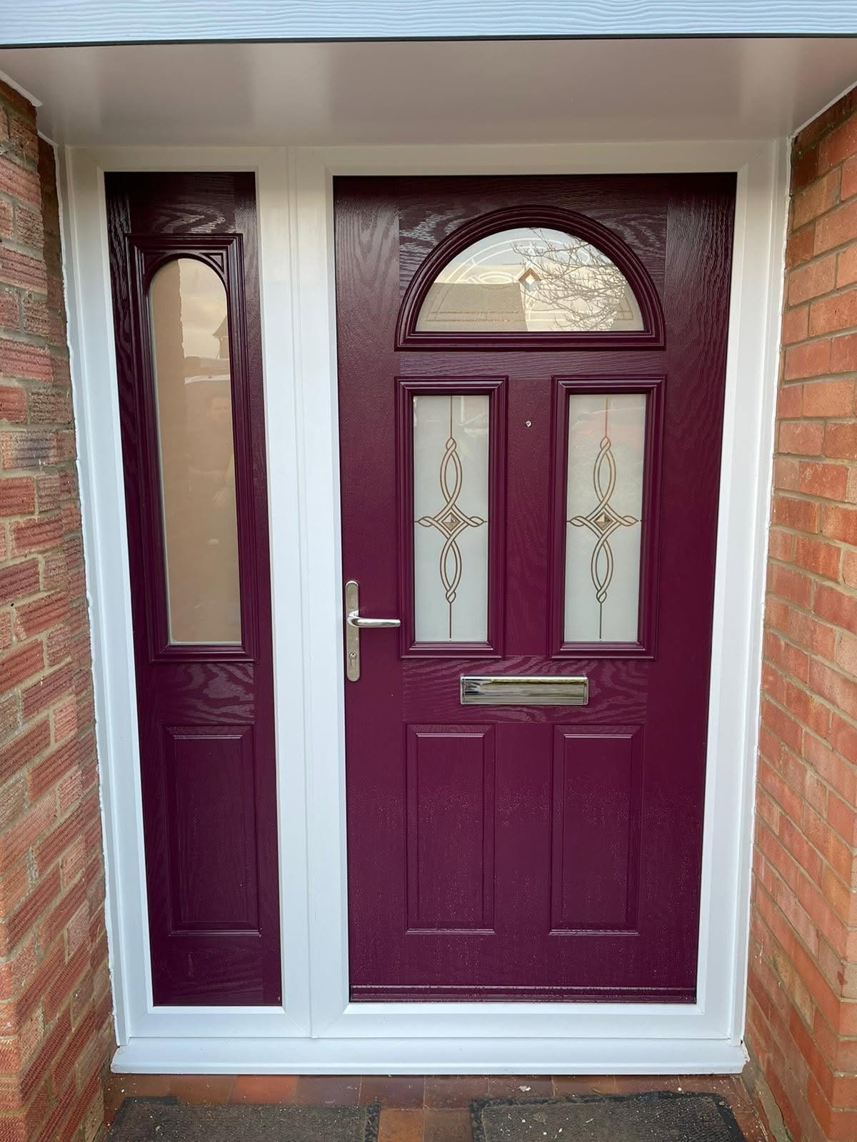 A maroon front door with decorative glass panels and a mail slot, flanked by side glass panels with matching decorative glass, set in a brick wall.