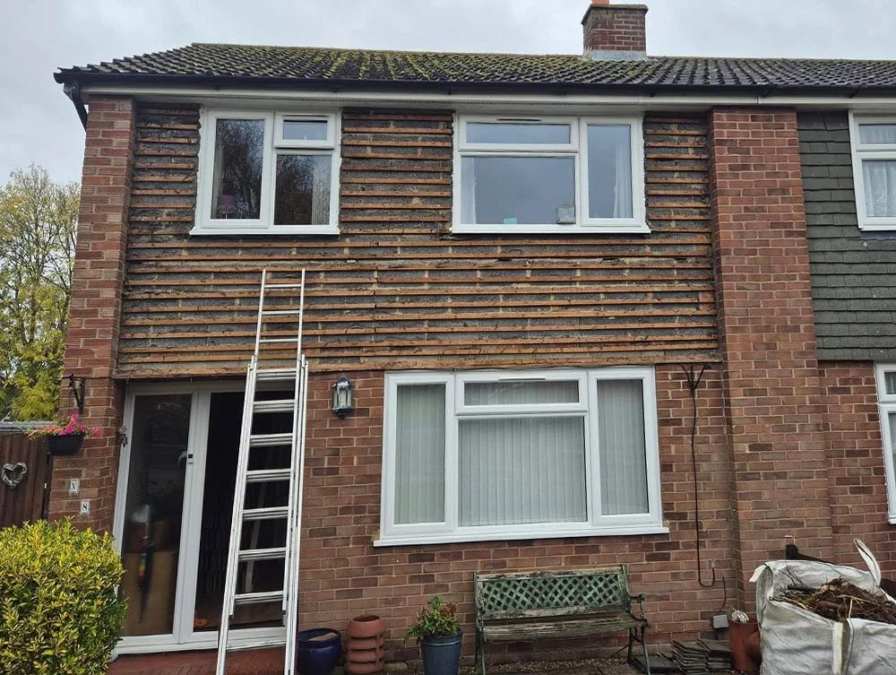 The exterior of a brick house undergoing window replacement or renovation, with a ladder leaning against the upper part of the house, and a garden bench, plants, and gardening supplies in the front yard.