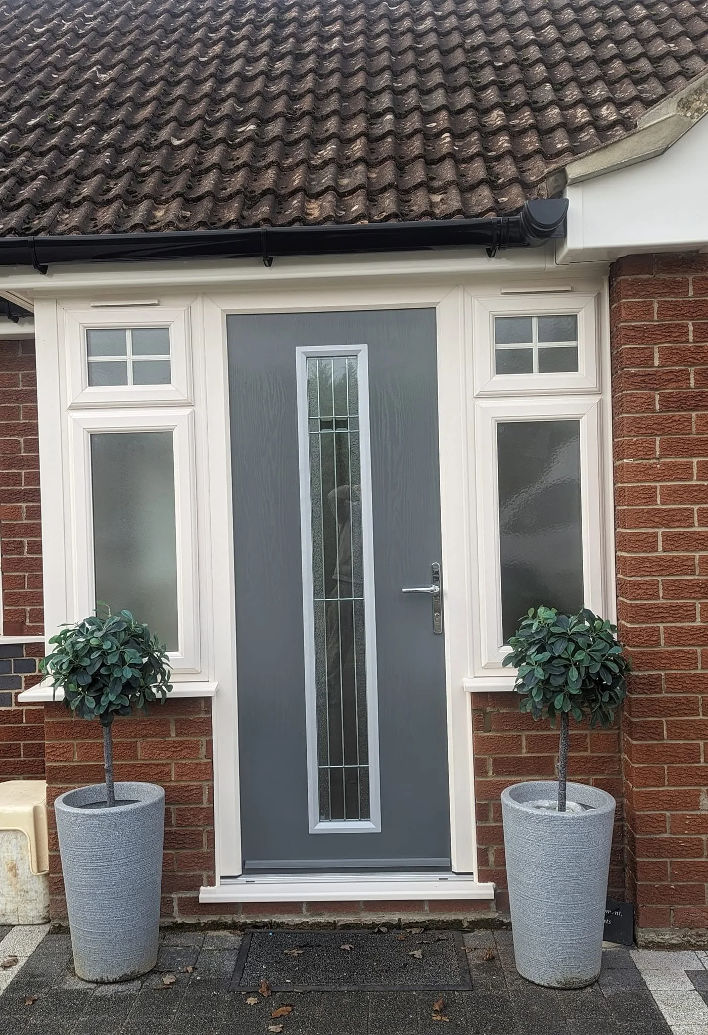 Front door of a house with a brick exterior, white trim around the door, and two potted trees on either side of the door.
