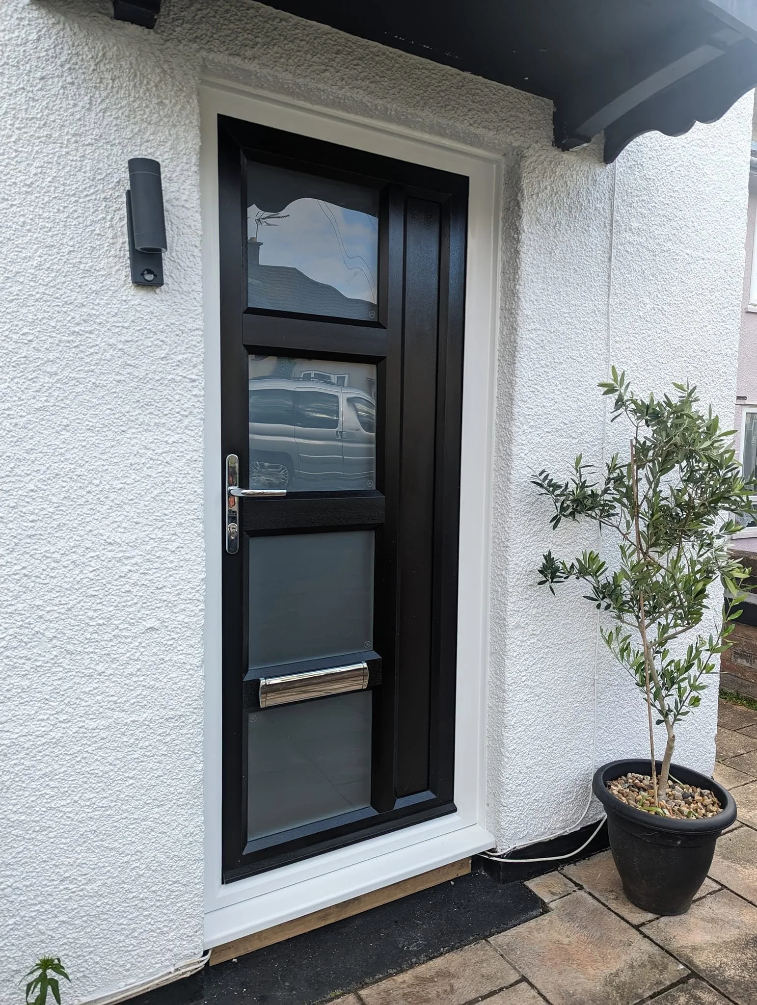 A black front door with glass panels installed in a white textured exterior wall, with a black handle and lock, next to a wall-mounted light fixture. A potted small tree with green leaves and a gravel top in a black pot is positioned to the right of 