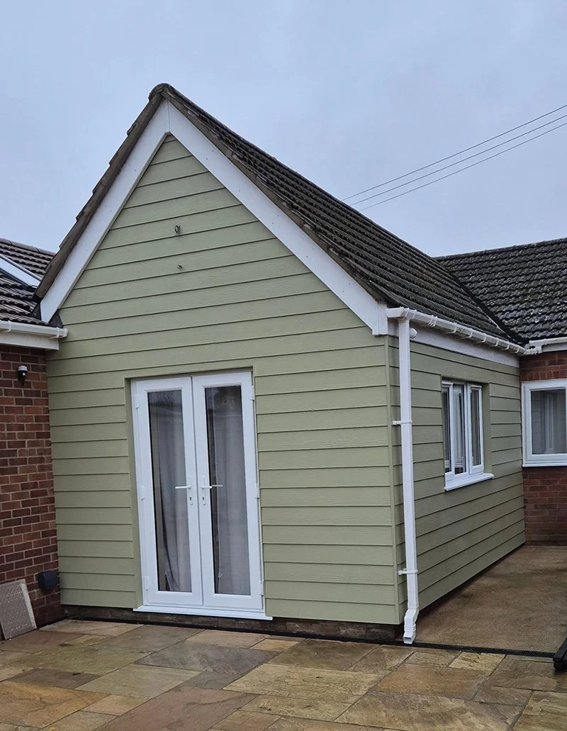 A small extension with beige siding and white framed double glass doors, attached to a brick house, under a gray sky.