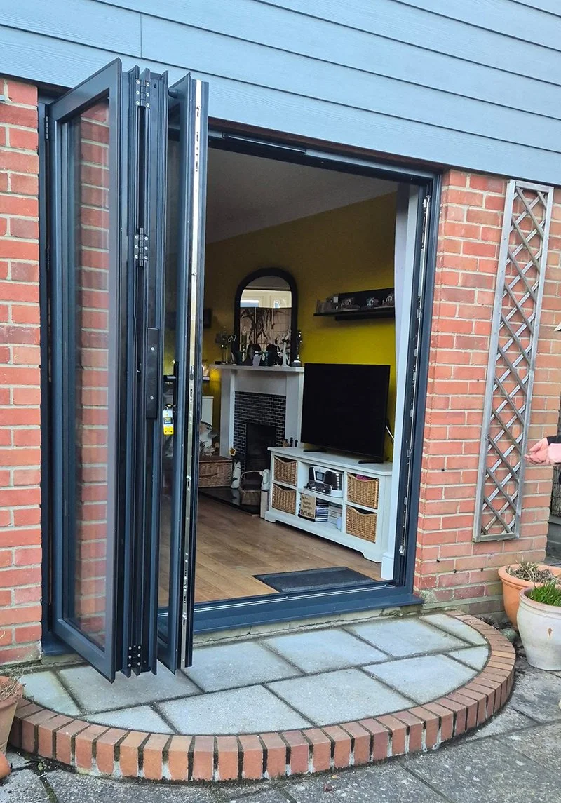 Open glass door leading into a home with a brick and blue siding exterior, showing a living room with a TV, fireplace, and yellow wall.