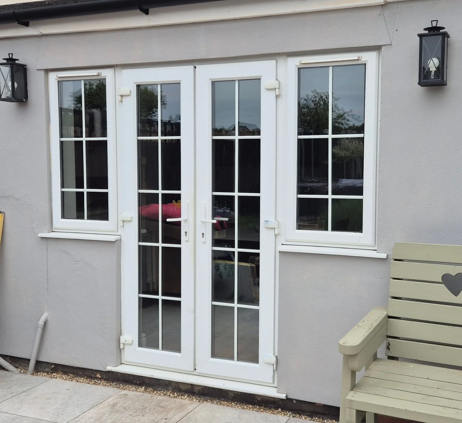 Exterior view of a house with white French doors and windows, gray stucco walls, and black outdoor wall lanterns on either side of the doors. A beige bench is on the right side of the image.
