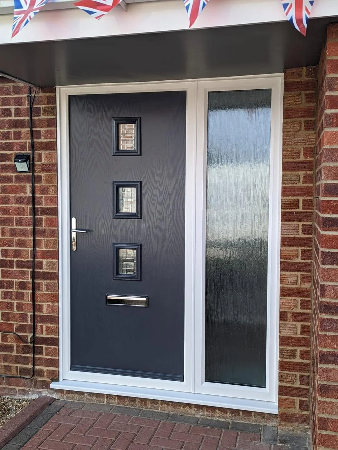 A modern front door with three small windows arranged vertically, a silver mail slot, and a silver handle, next to a textured glass side panel, all set within a brick exterior.