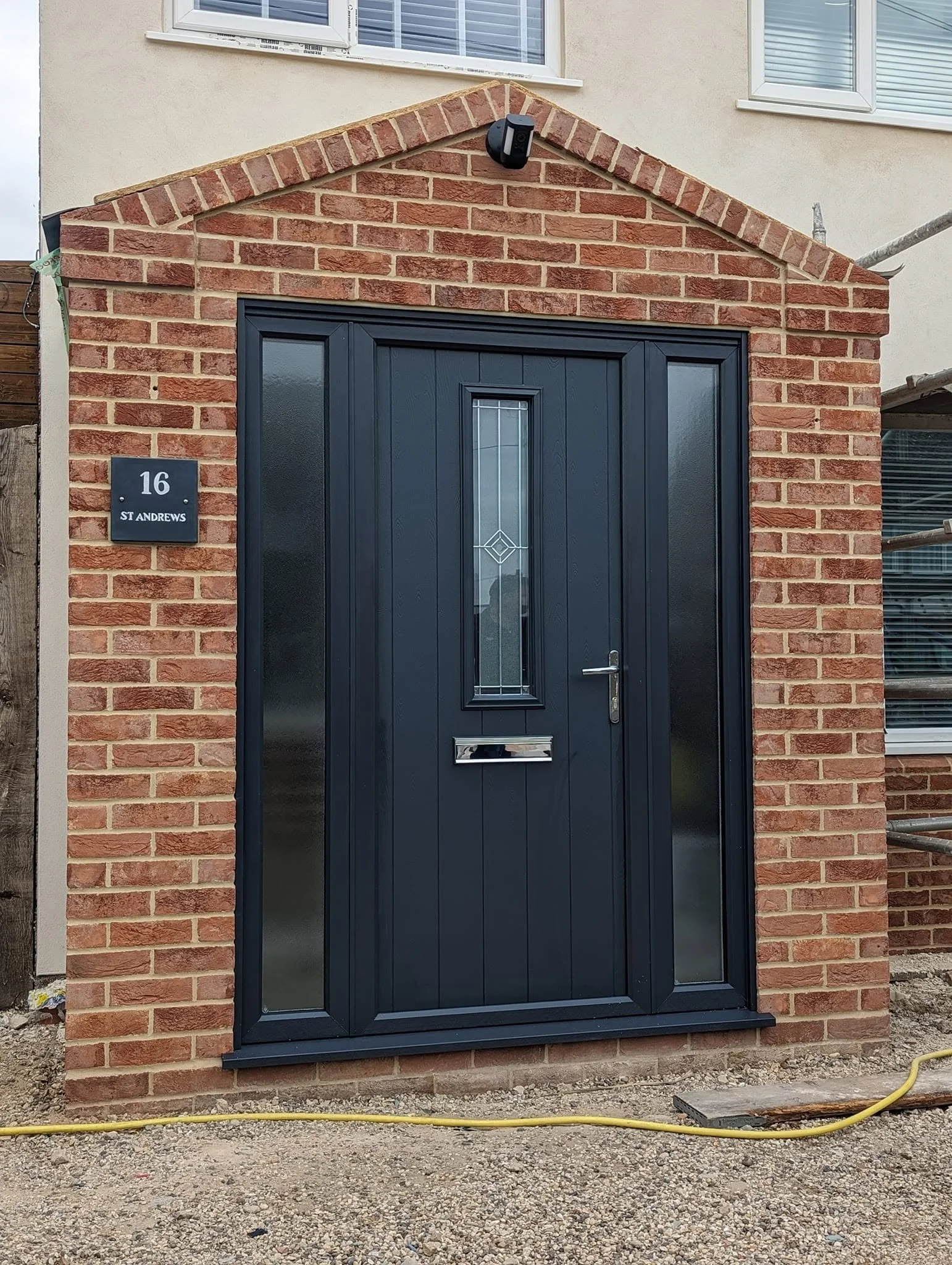 New black front door with small window and mail slot, surrounded by red brick wall, with house number 16 and street name 'St Andrews' on a plaque, and security camera above the door, with some construction materials and gravel in front.