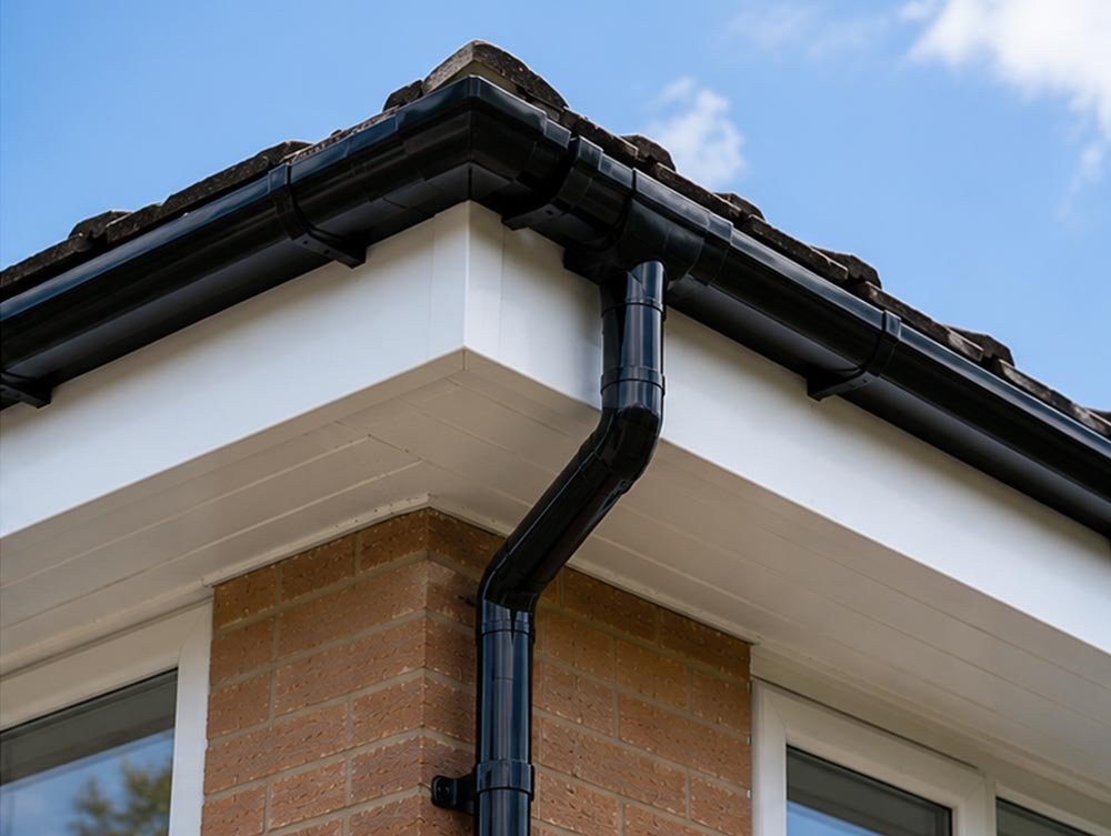 Close-up of a house corner with a black downspout, roof tiles, and a brick wall against a blue sky