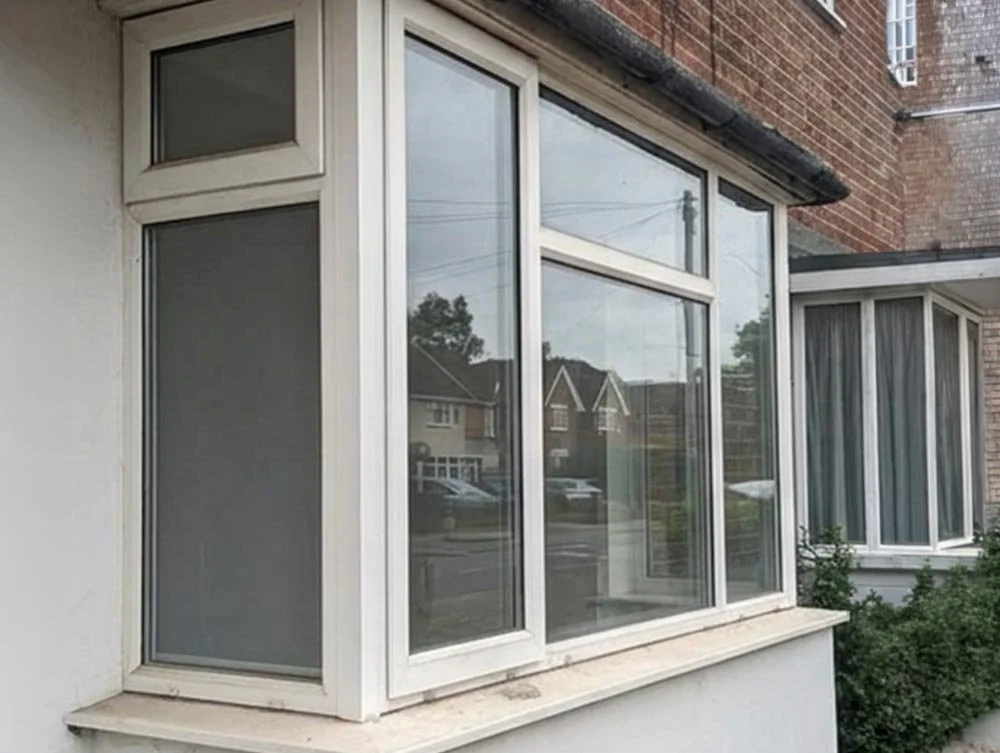 Close-up view of a large bay window on the side of a house with white framing, showing reflections of neighboring houses and trees.