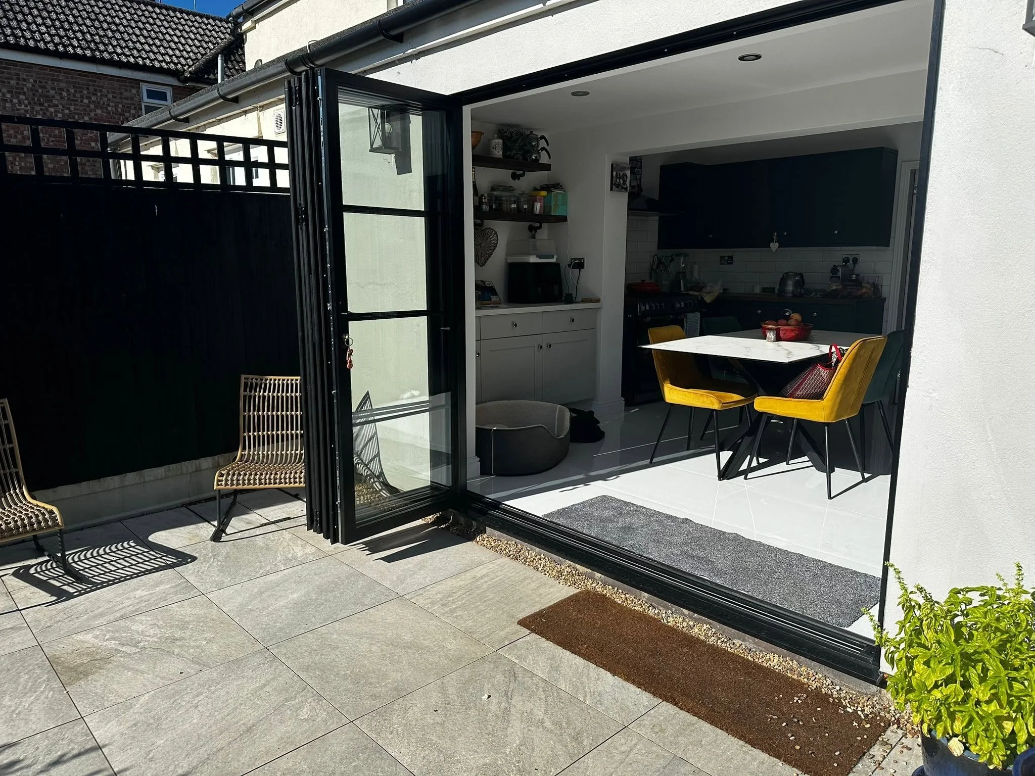 A backyard patio with gray tile flooring, a black metal fence, and an open glass door leading to a kitchen. Outside, there are two woven chairs and a small table. Inside, the kitchen has dark cabinets, a white countertop, and various appliances. Ther