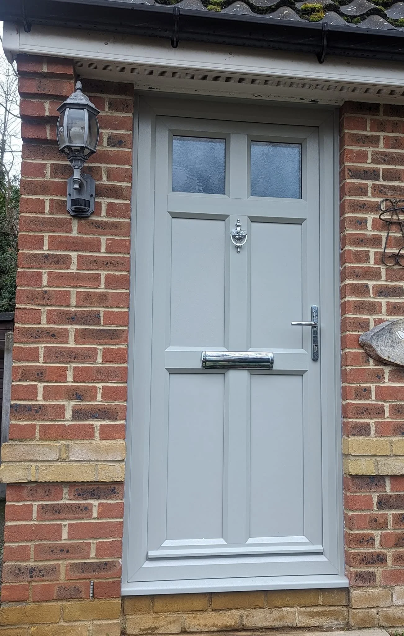 A gray front door with a shiny mailbox in the middle, set in a brick wall with a lantern on the left side, and a decorative metal piece on the right side.
