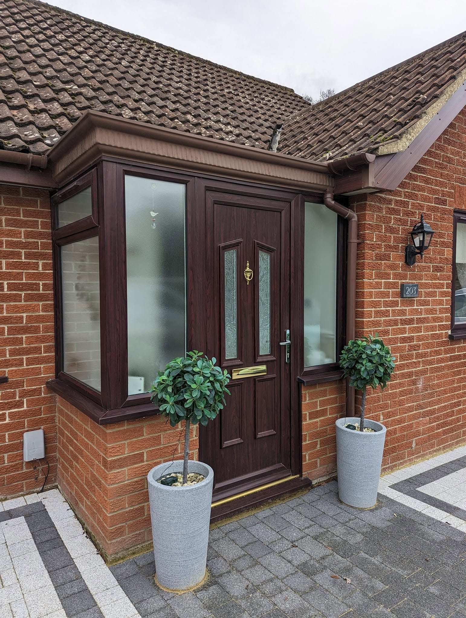 The image shows the front door of a house with a brick exterior. There are two tall potted plants with green foliage on either side of the door. The door is brown with frosted glass panels and a gold handle. To the right of the door, there is a black
