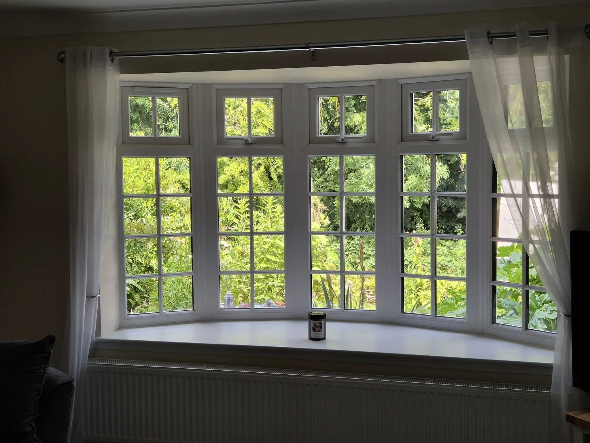 A large bay window with multiple small panes, flanked by white curtains, looking out onto lush green trees and plants outside.
