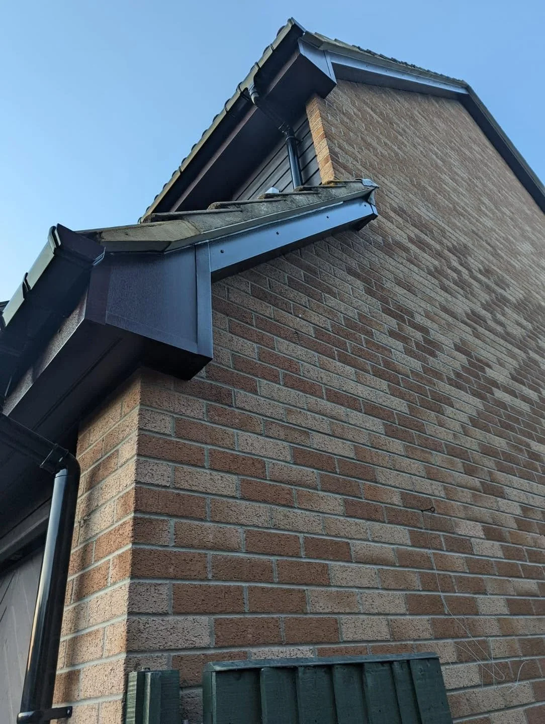 Close-up view of the corner of a brick house roof with black rain gutters and a chimney against a clear blue sky.