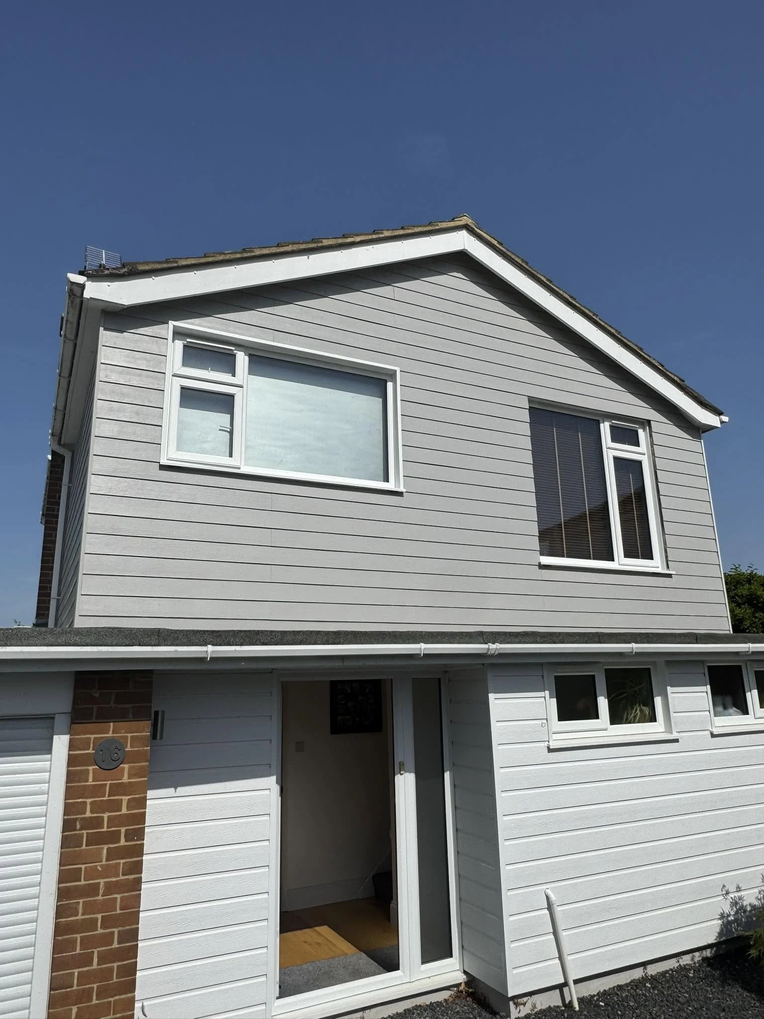 A modern house with gray horizontal siding, white window frames, a brick accent on the left, and a garage door at the bottom. The front door is open, revealing a small entryway with a rug. The house has a sloped roof with shingles, and part of a chai