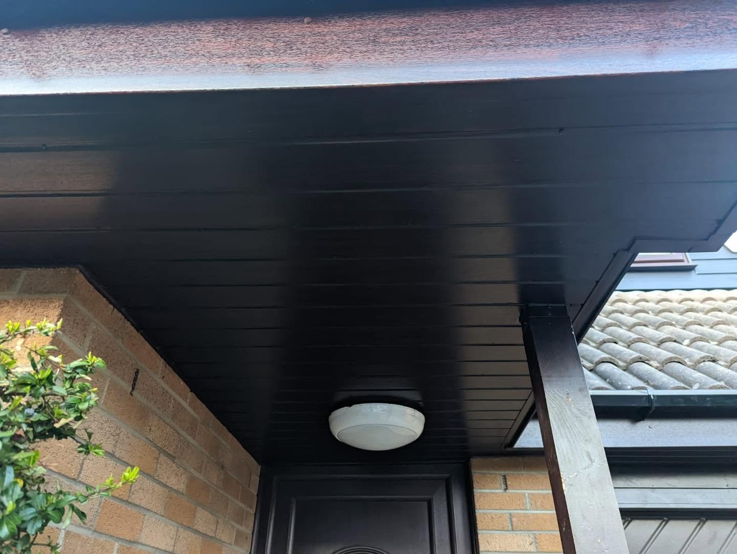 Overhang of a house with black siding, a white ceiling light fixture, a brick wall, and a corner of a tile roof with grey shingles.