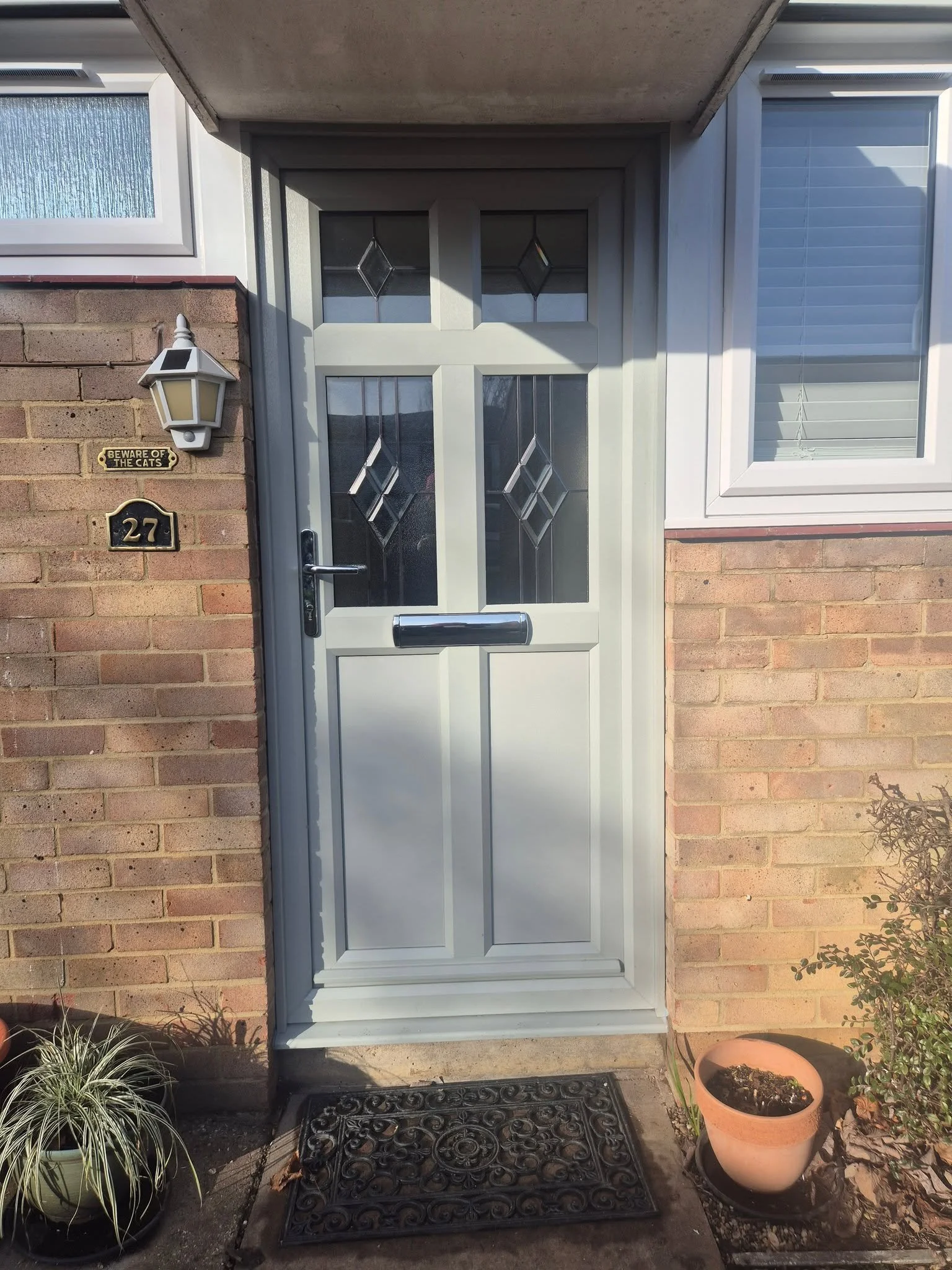 A front door with glass panels on a brick house. There is a lamp and house number '27' on the left, along with a sign saying 'Beware of the Cats'. Potted plants and a doormat are on the porch.